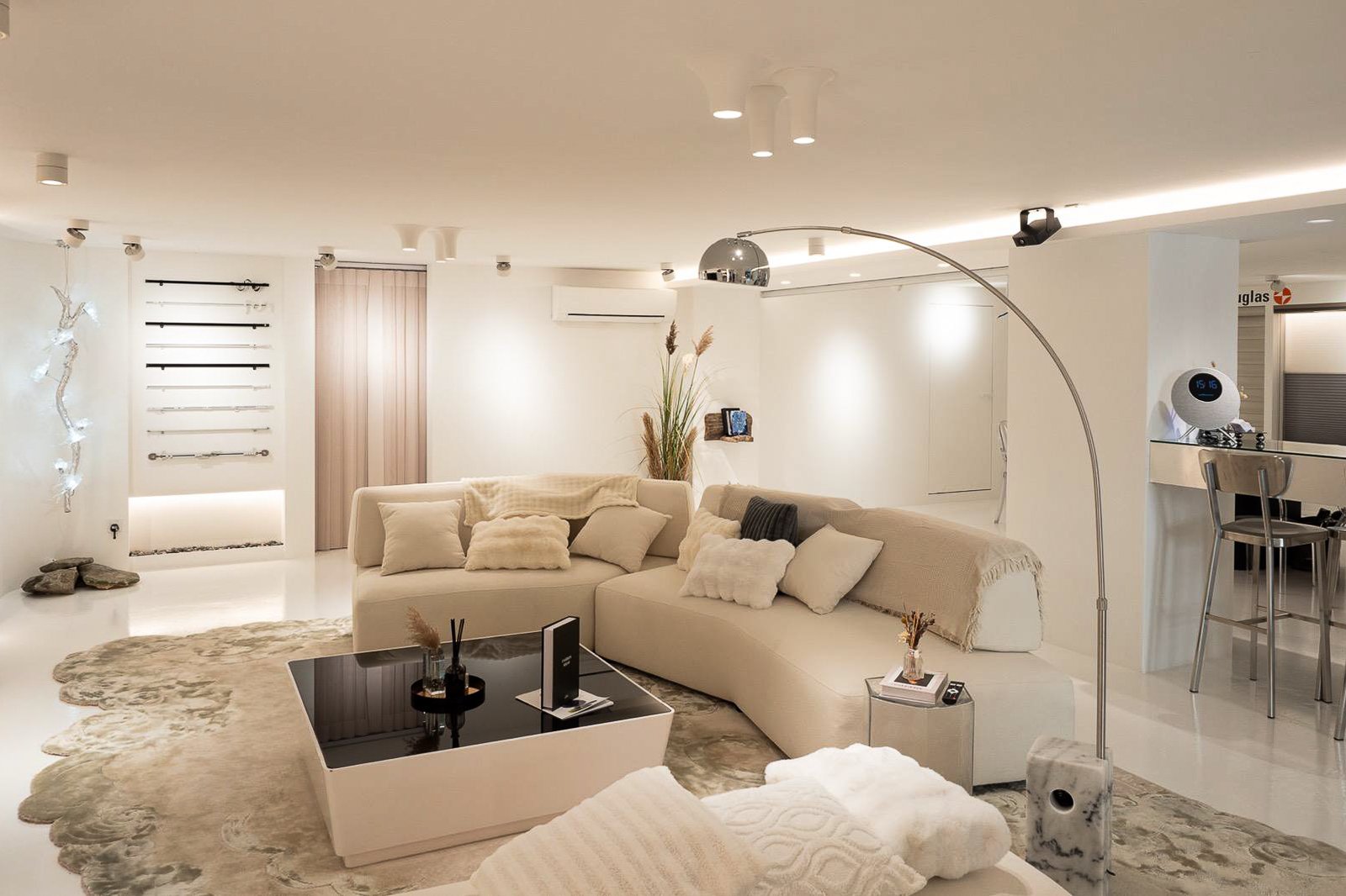 Modern living room with beige sectional couch, pillows, and a black coffee table. Decor includes wall-mounted bars, a large floor lamp, and decorative plants. The room is well-lit with a white and neutral color scheme, and a small bar area is visible in the background.