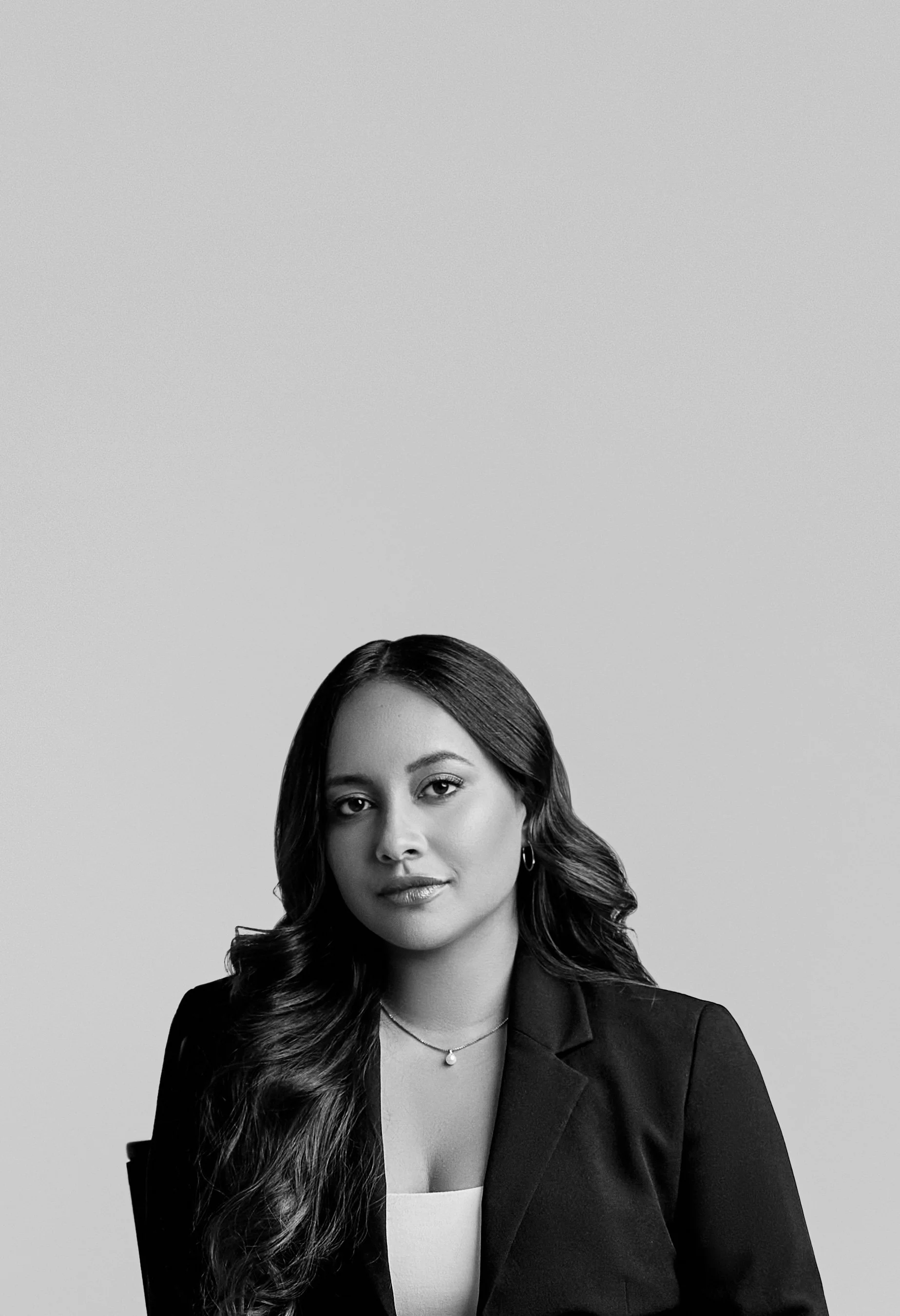 Black and white portrait of a woman with long wavy hair, wearing a blazer, necklace, and hoop earrings, looking at the camera with a confident expression.