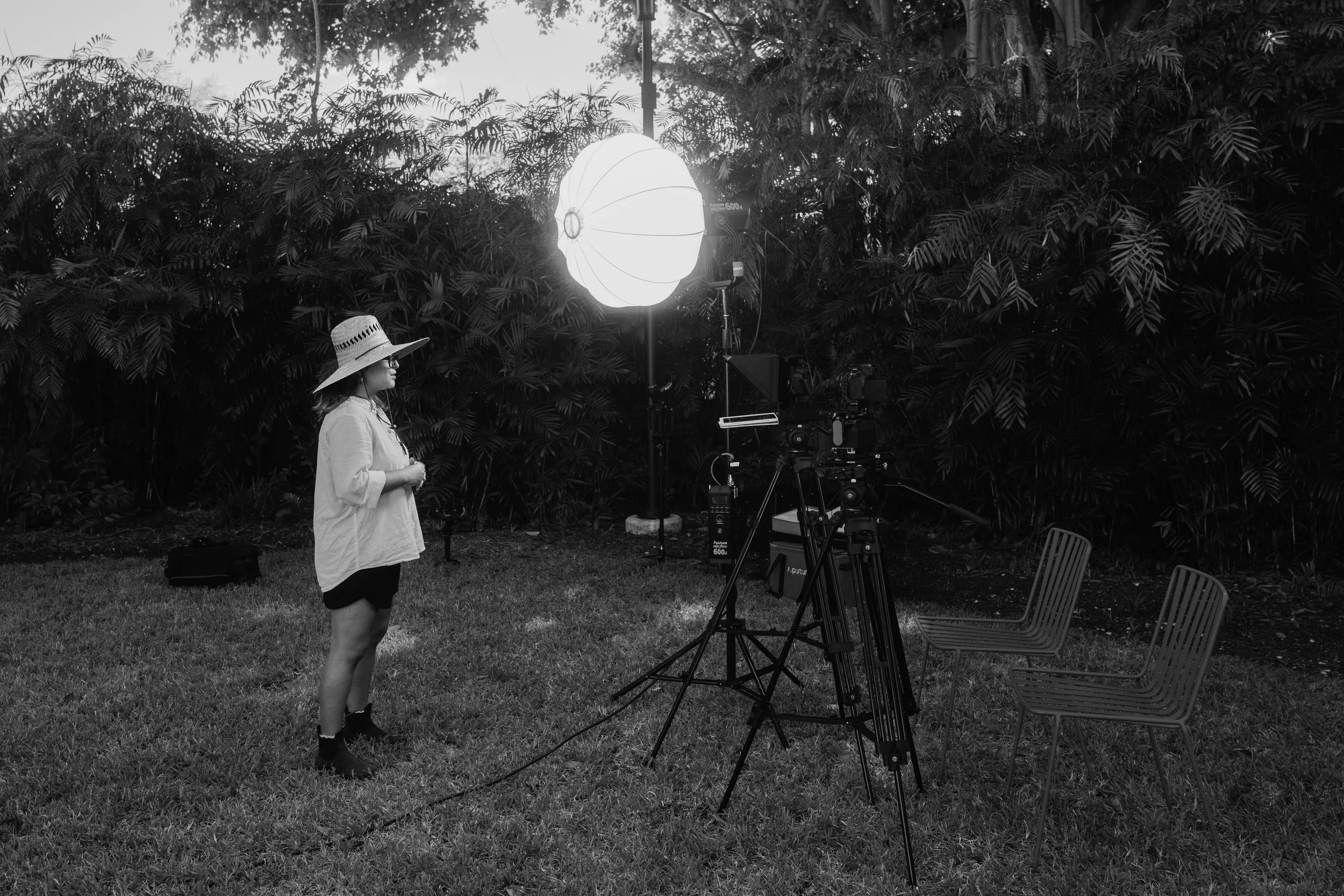 A woman in a large sun hat stands outdoors at a filmmaking set with professional lighting and camera equipment, surrounded by dense foliage.