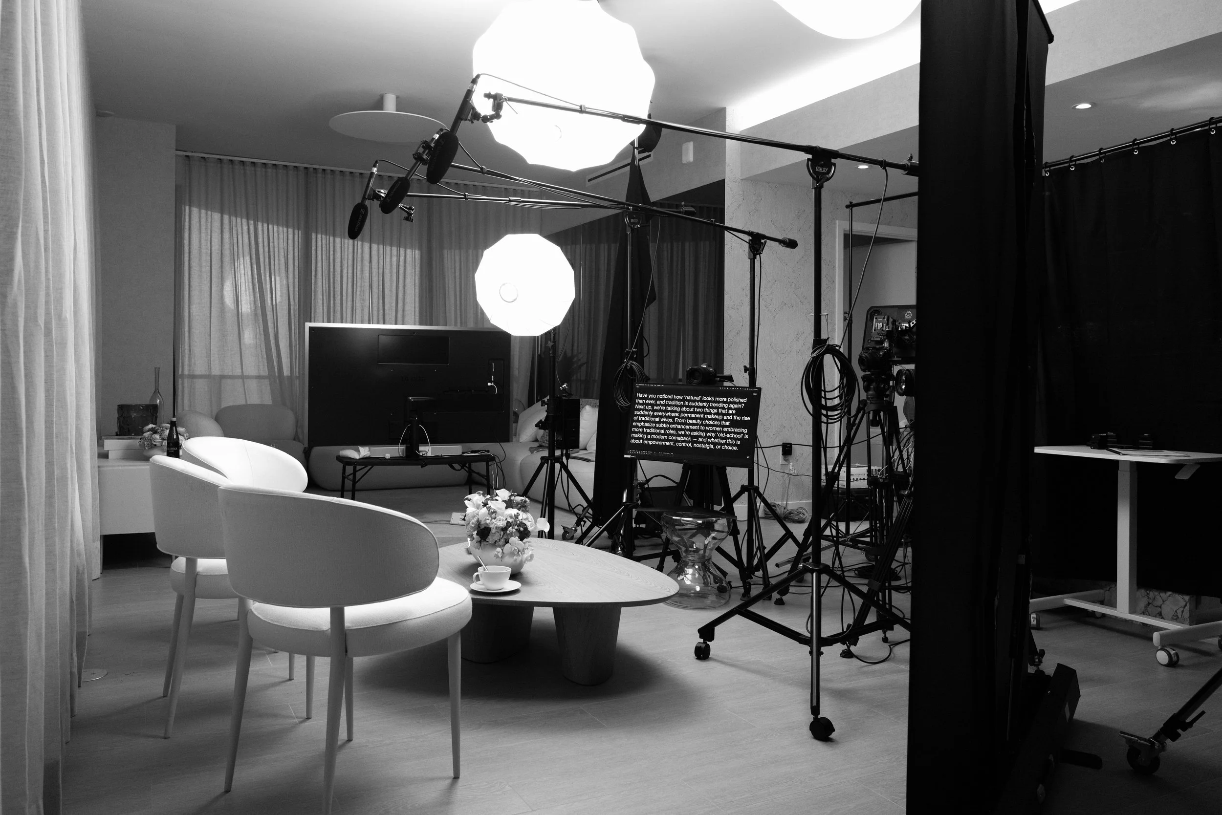A behind-the-scenes view of a television or film production set, with lighting equipment, cameras, and microphones positioned around a scene that features a round table with a flower arrangement, cups, and a seating area.