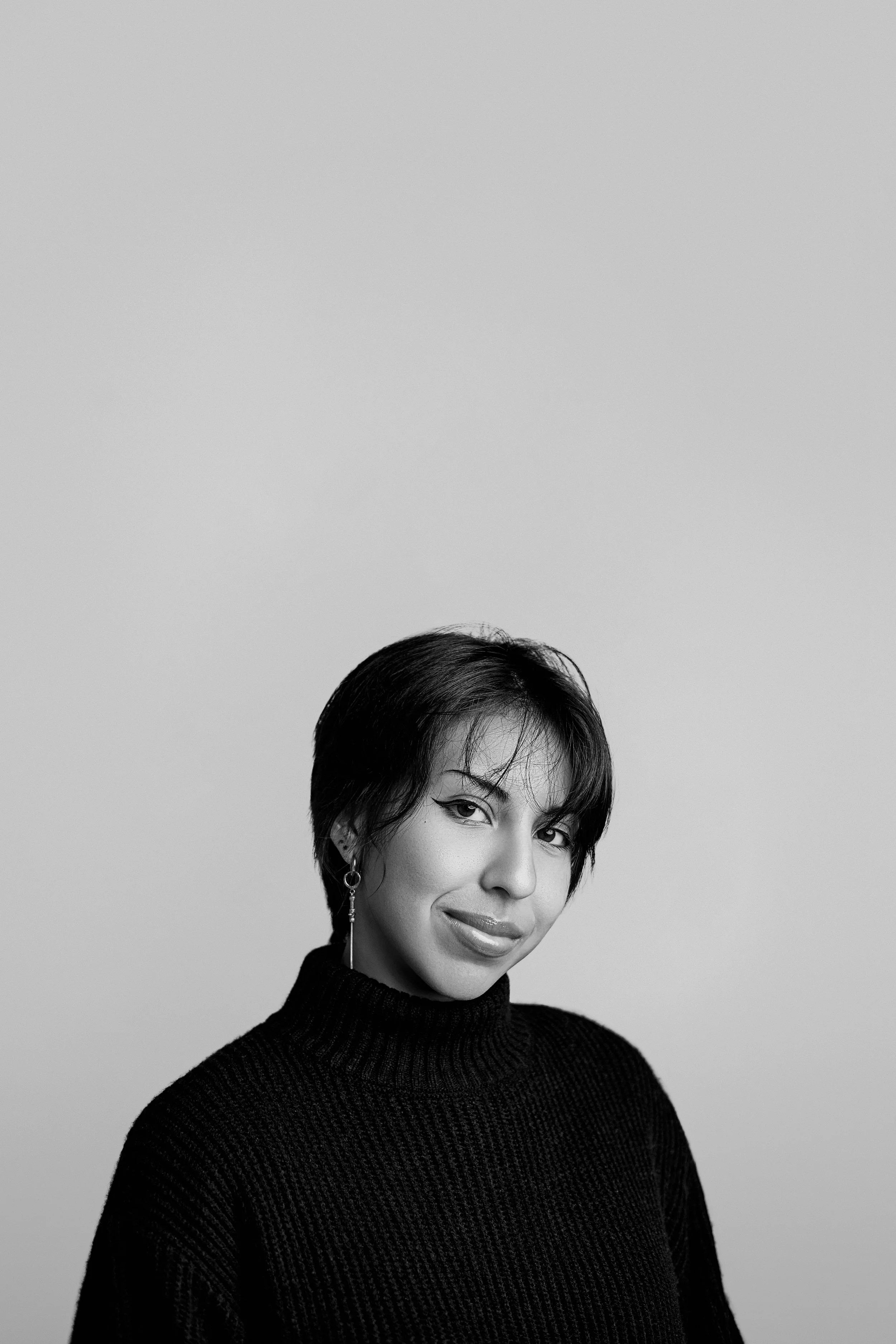 A black and white portrait of a woman with short, dark hair, wearing earrings and a high-neck sweater, looking at the camera with a slight smile, against a plain light background.