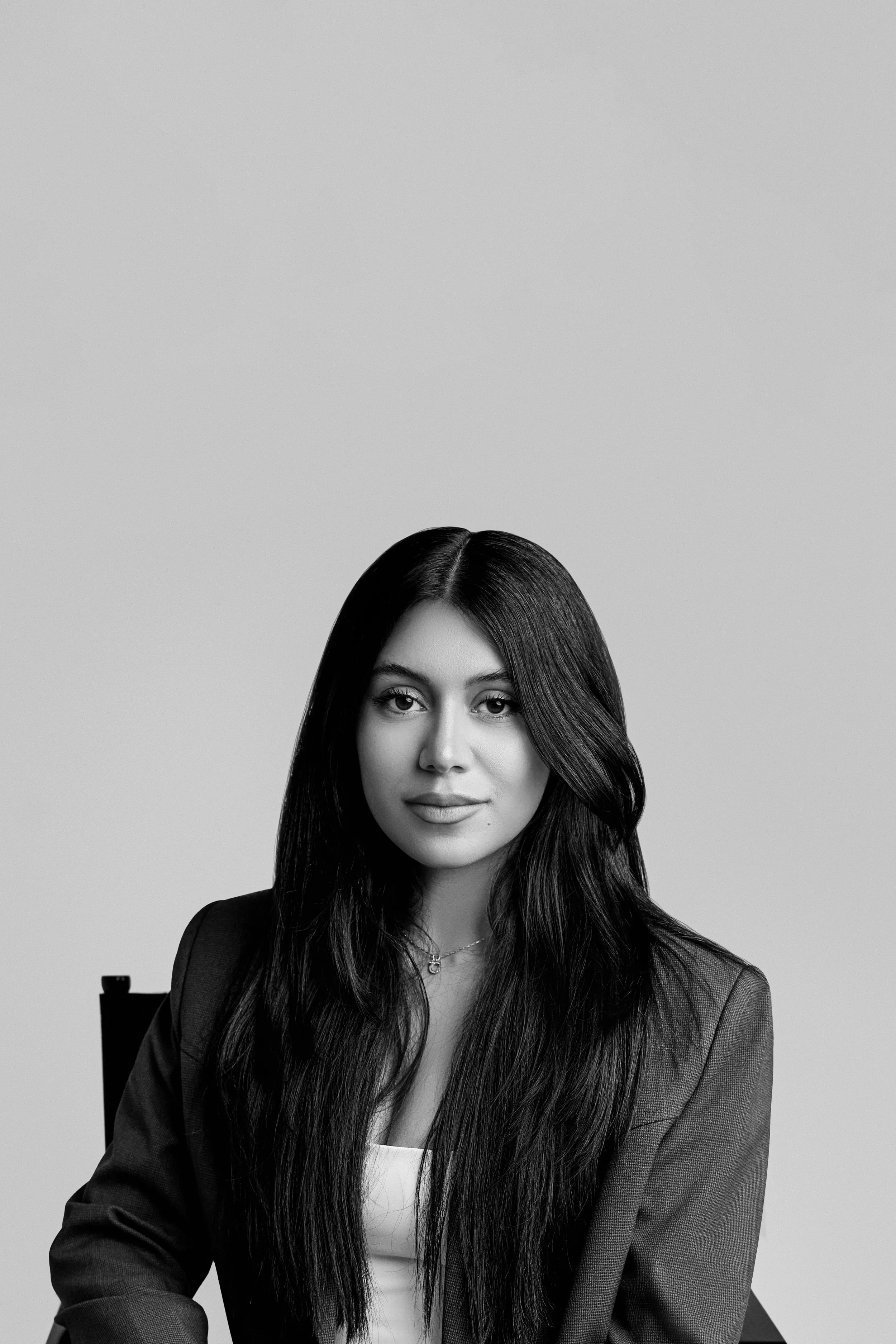 A young woman with long, straight, dark hair and light skin, wearing a blazer, sitting on a chair against a plain, light background.