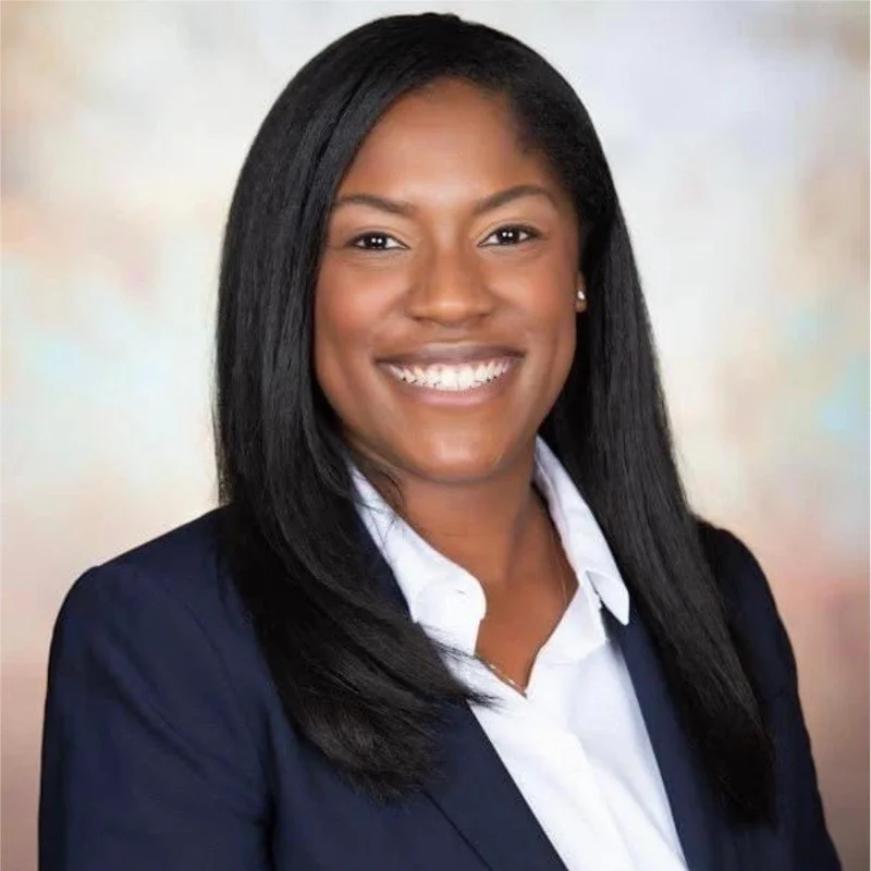 A professional woman with long black hair, wearing a navy blazer and white shirt, smiling at the camera.