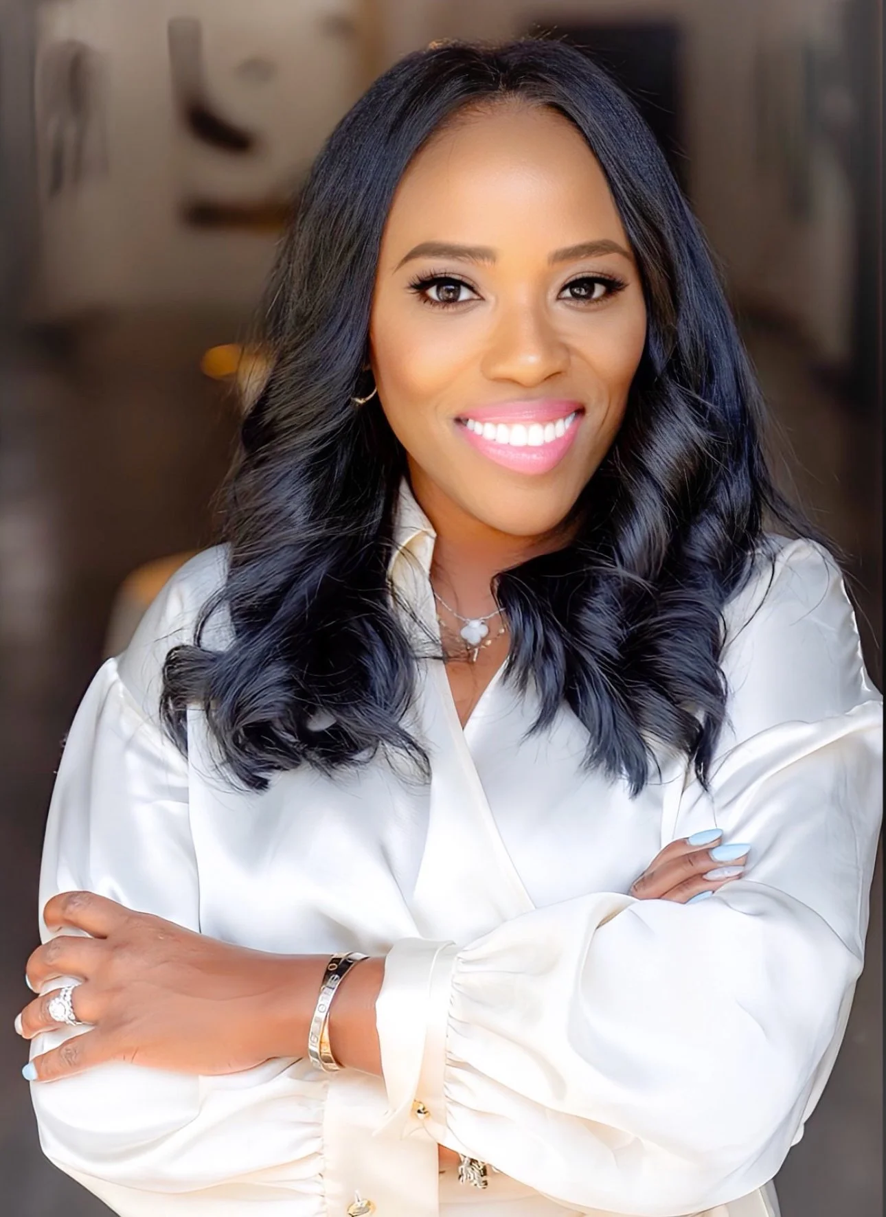 A woman with long, dark wavy hair smiling at the camera, wearing a white satin blouse and jewelry, in an indoor setting.