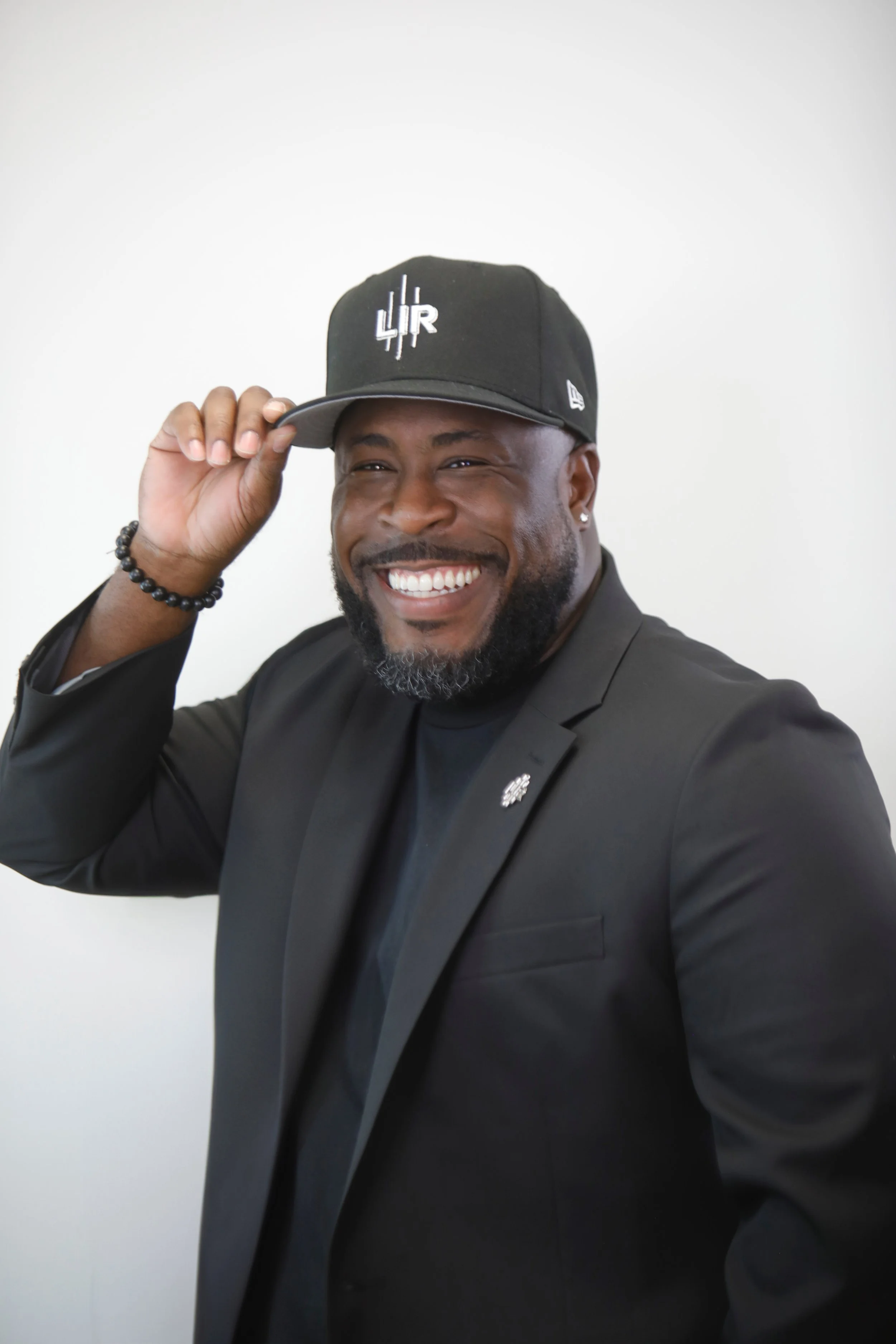 Smiling man in a black suit, wearing a black cap with white lettering, adjusting the cap with his right hand, against a plain white background.