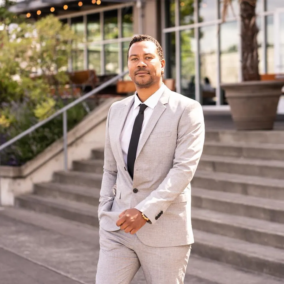A man in a light-colored suit with a white shirt and black tie standing outdoors on steps with a building and some greenery in the background.
