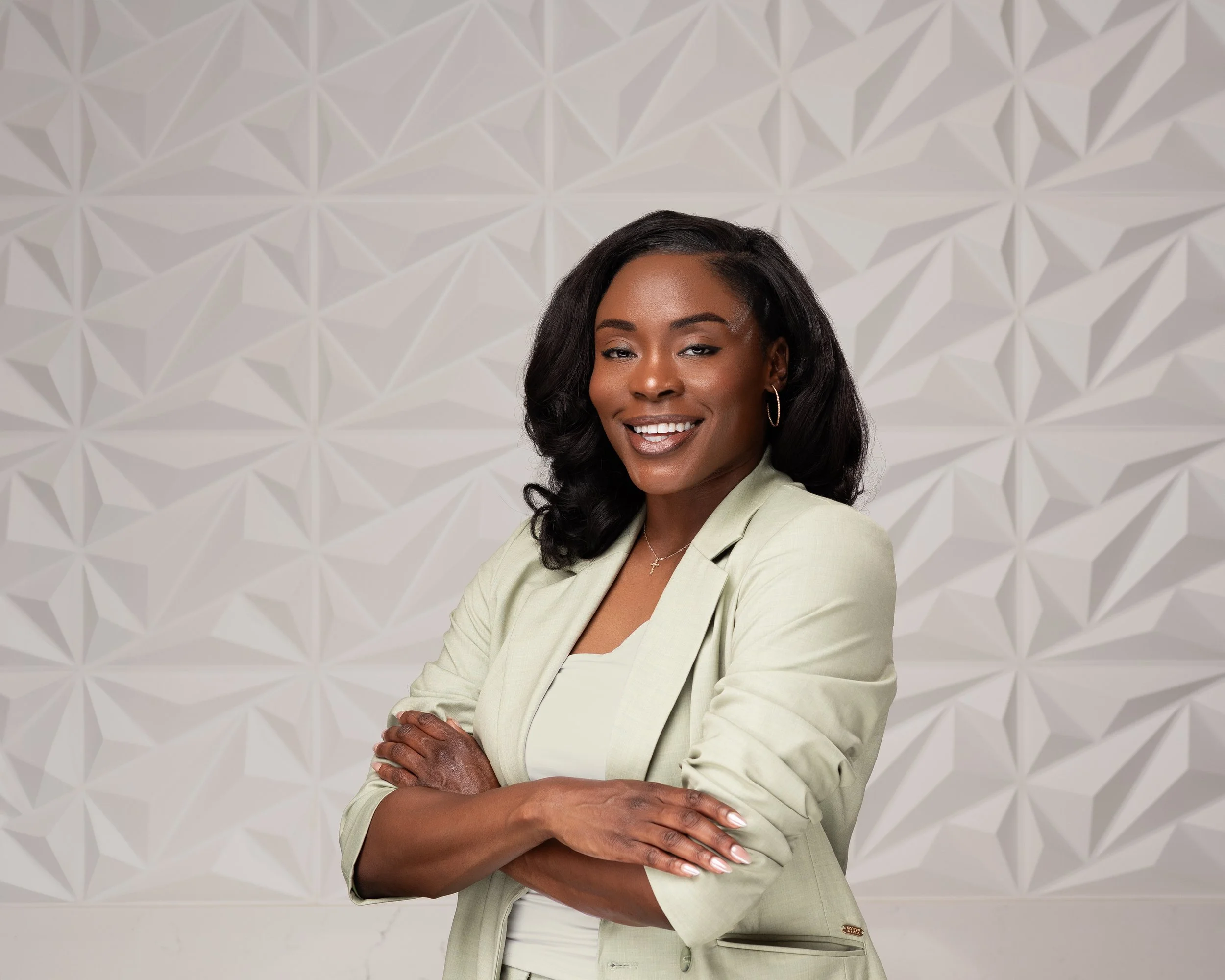 A confident woman with dark hair styled in loose curls, wearing a light-colored blazer and top, smiling with arms crossed, standing in front of a textured geometric wall background.