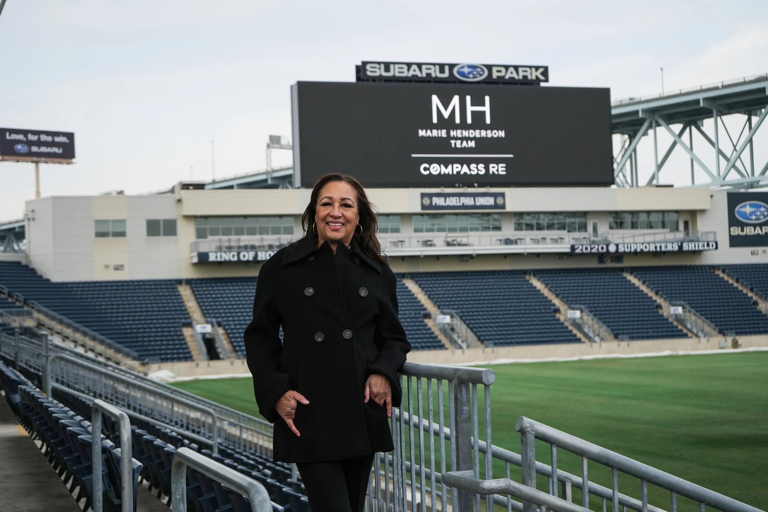 A woman standing in a sports stadium with an electronic scoreboard behind her displaying 'MH Marie Henderson Team Compass RE'. The stadium seats are visible, and the field is green with a bleacher in the foreground.