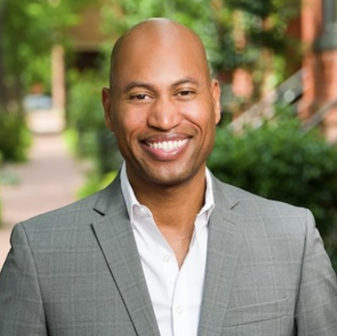 A smiling man in a light gray suit with a white shirt stands outdoors in a green, leafy area.