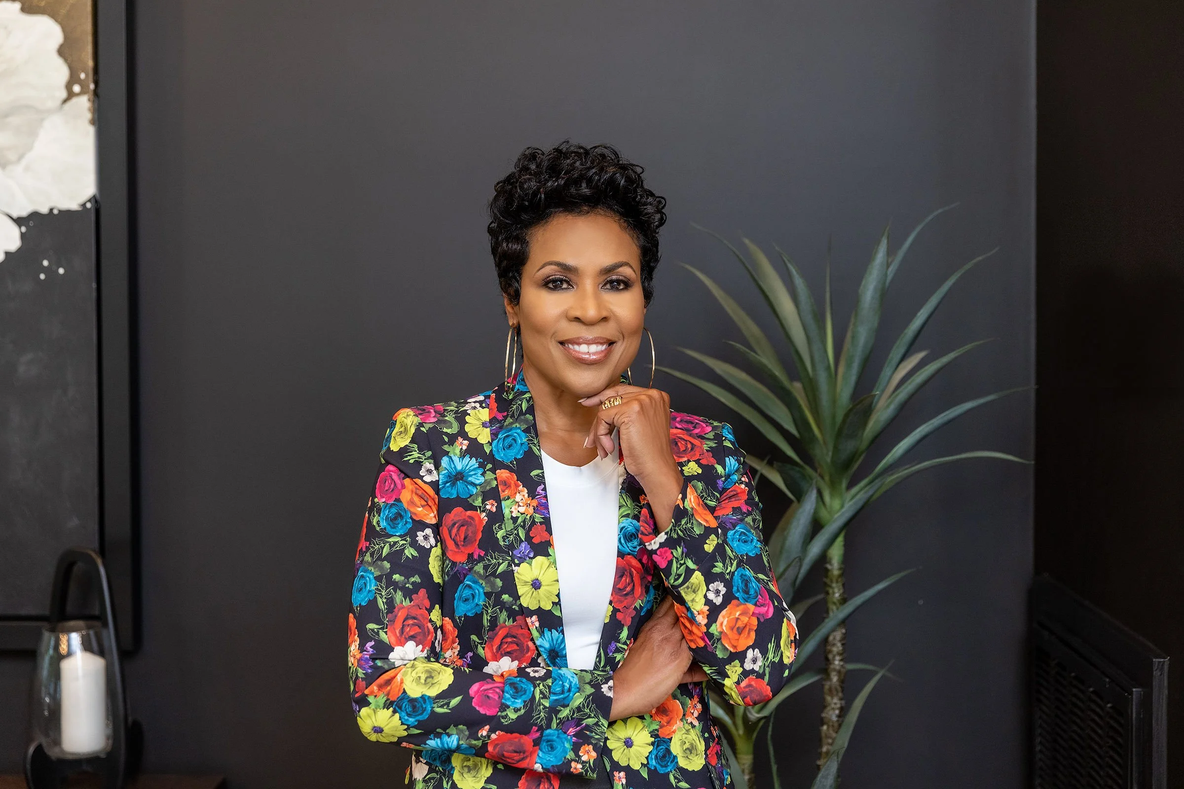 A woman with short curly black hair smiling, wearing a colorful floral blazer and hoop earrings, standing with her arms crossed in front of a dark wall and a potted plant.