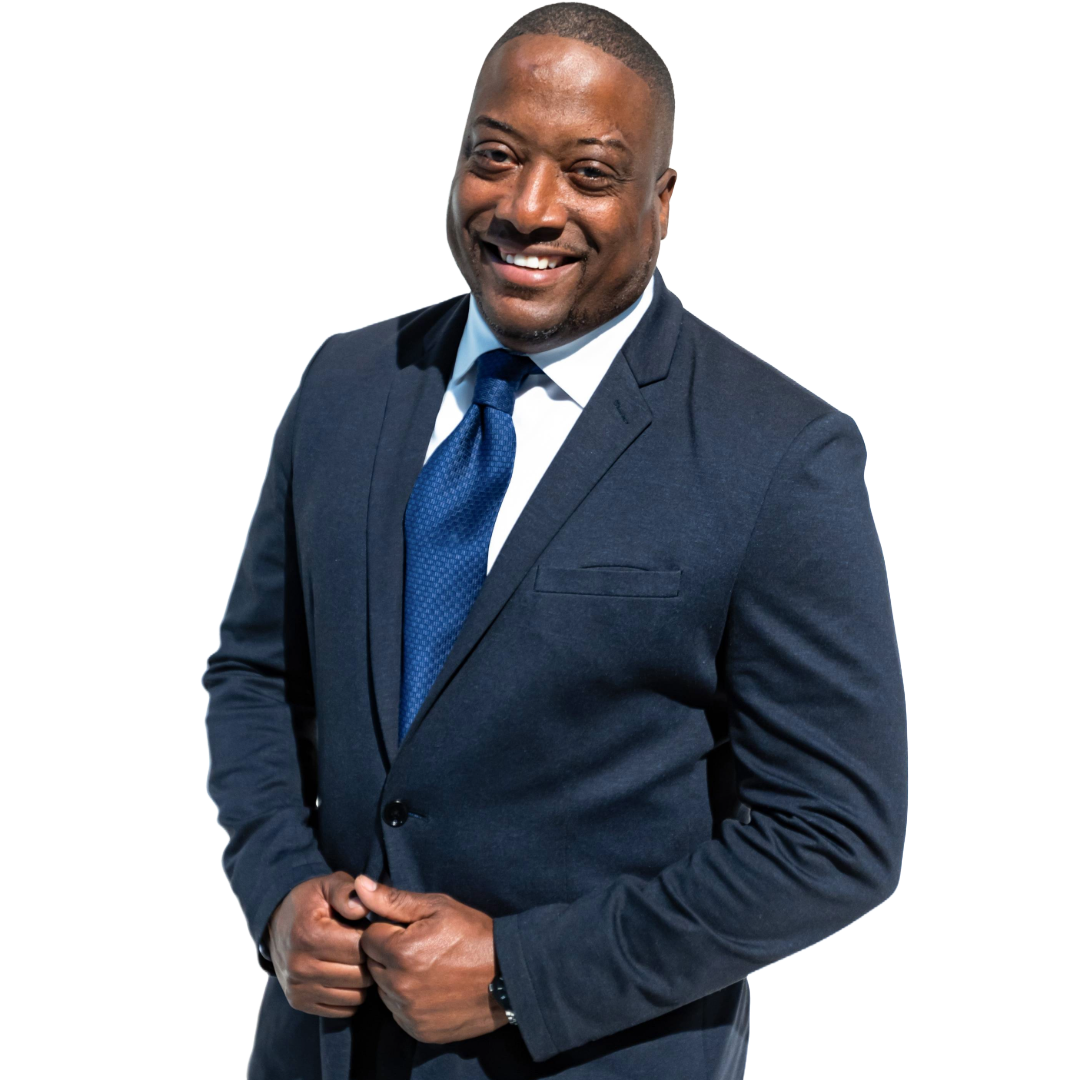 A confident African American man in a dark suit, white shirt, and blue tie, smiling and standing with hands clasped in front of a white background.