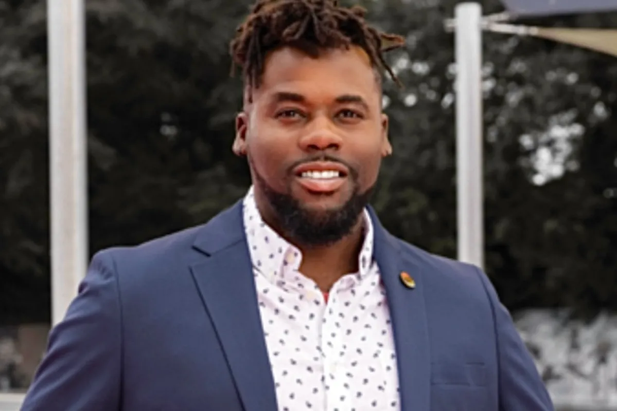 A smiling man with dreadlocks wearing a navy blazer and a white shirt with small polka dots, standing outdoors with trees and flagpoles in the background.