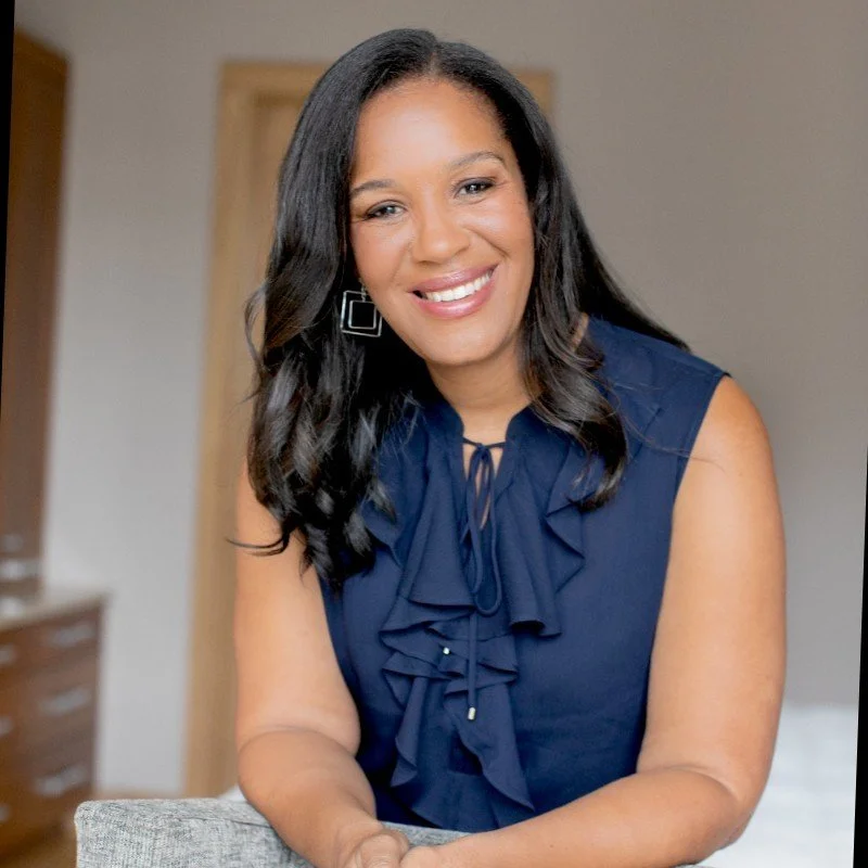 A woman with long dark hair wearing a navy blue sleeveless top with ruffles and square earrings, smiling and sitting at a table.