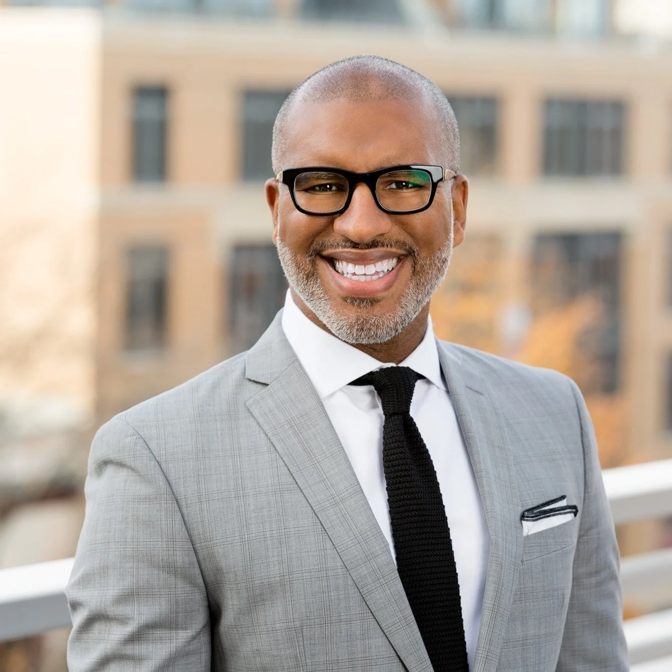A smiling man with glasses and a gray suit standing outside with a blurred cityscape background.