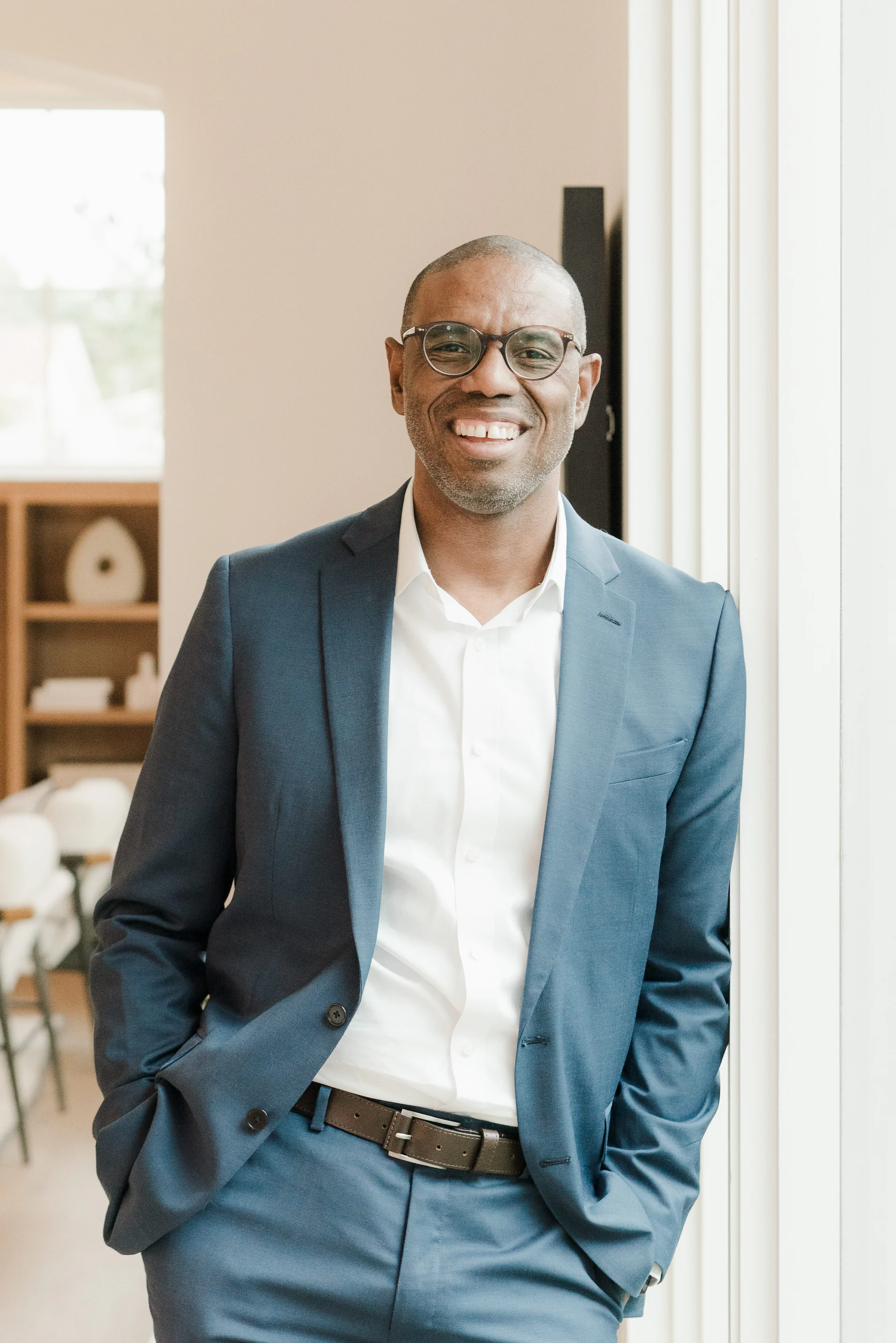 A man in a blue suit with a white shirt, wearing glasses, smiling and standing by a window.