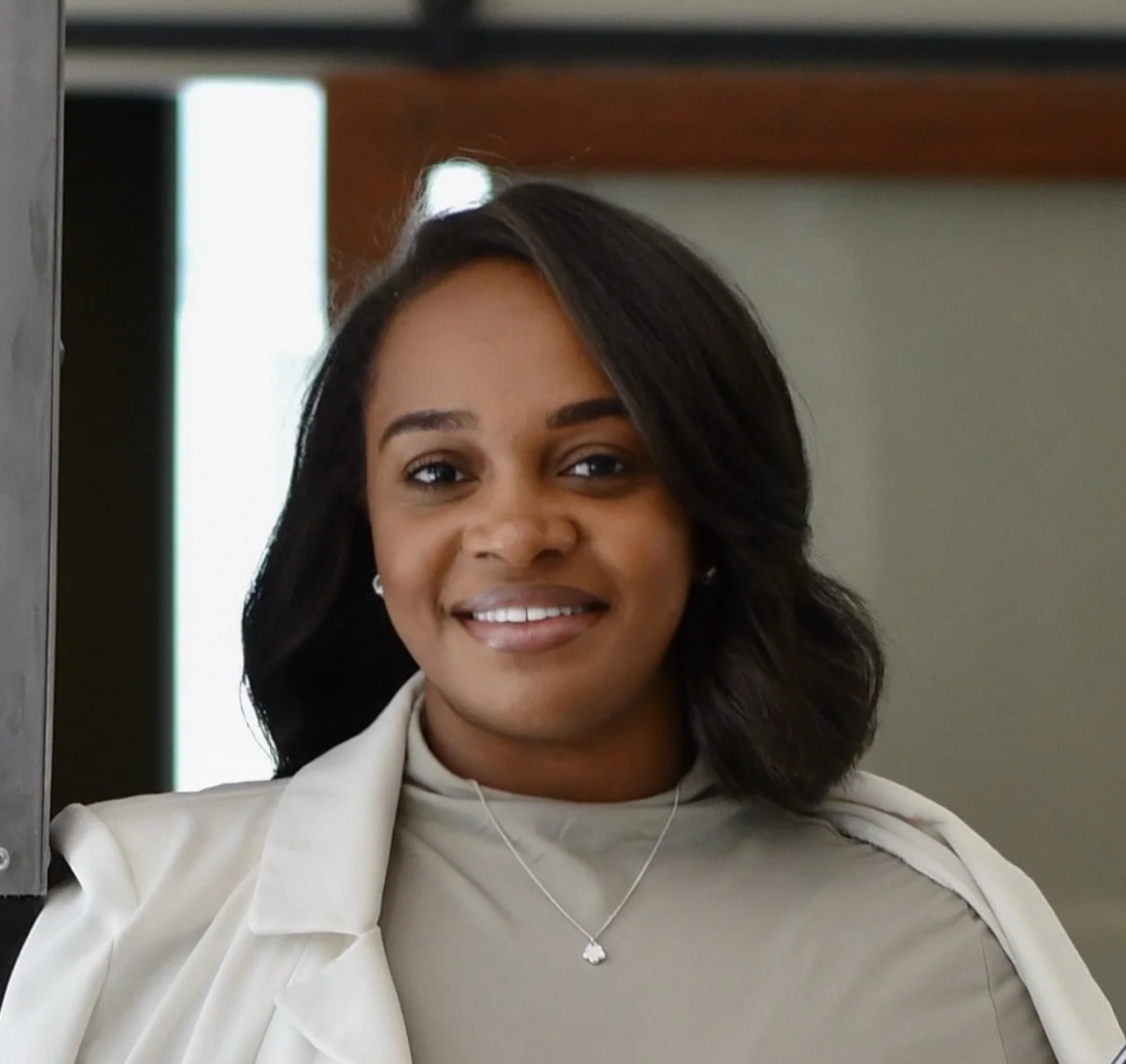 A woman with shoulder-length black hair, wearing a light-colored turtleneck and blazer, smiling in an indoor setting.