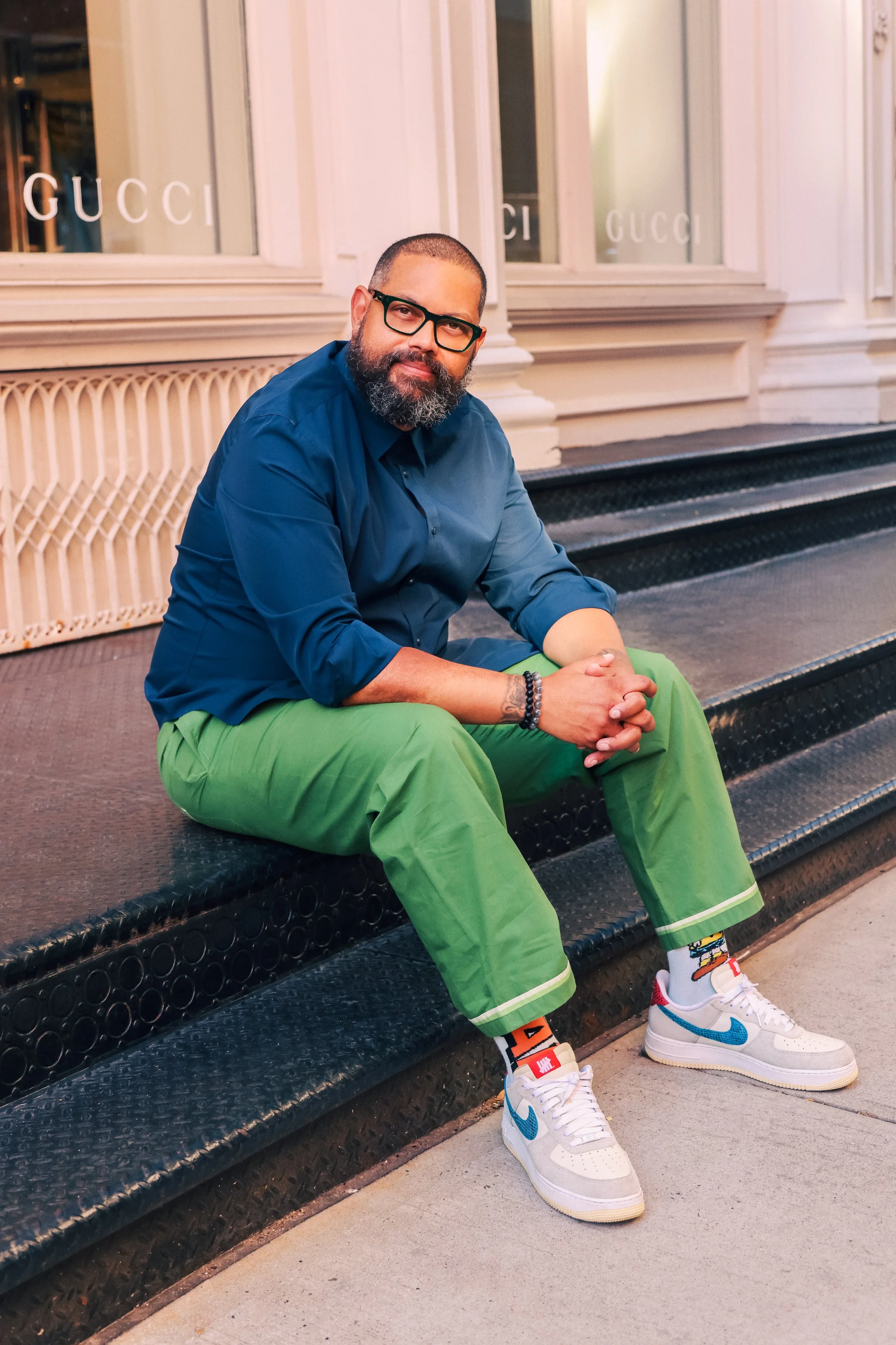 A man with glasses and a beard sitting on a black bench outside a building with 'Gucci' signs in the windows, wearing a blue shirt, green pants, and white sneakers.