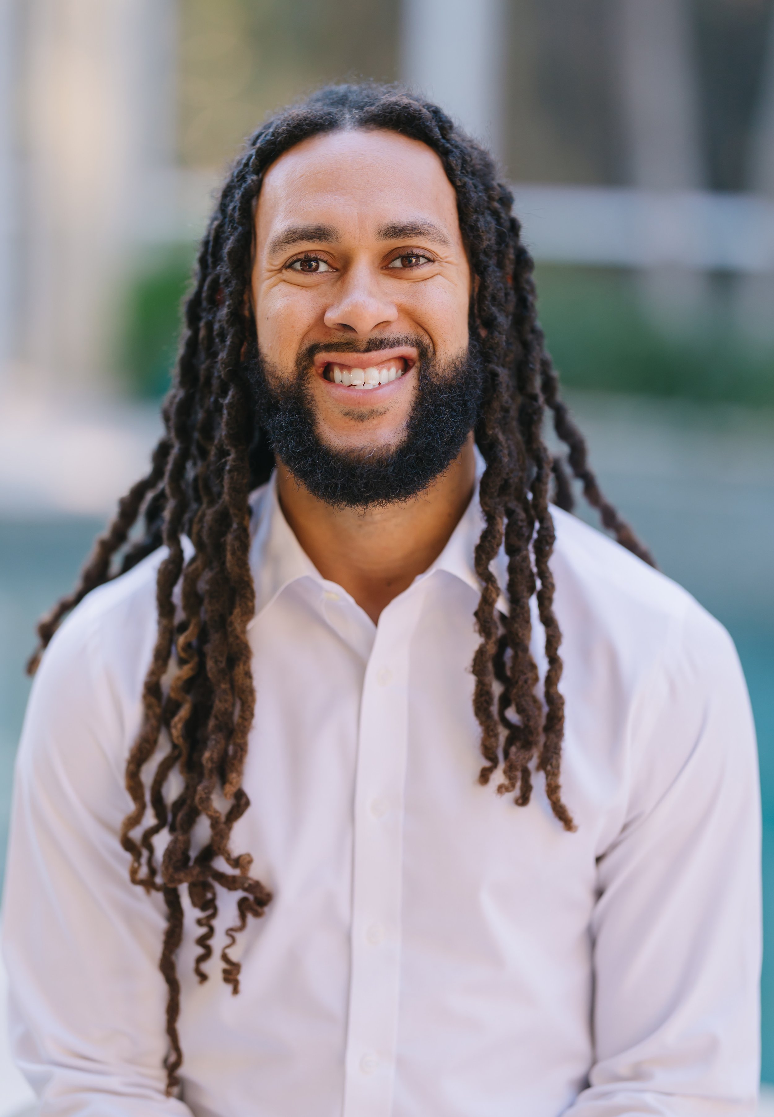 A man with long dreadlocks and a beard smiling, wearing a white button-up shirt, outdoors with blurred background.