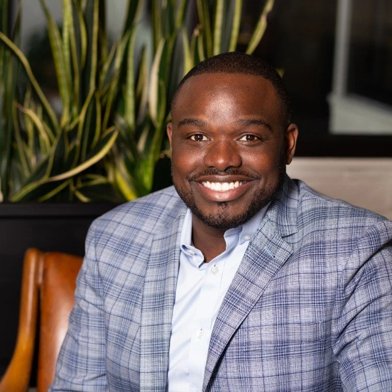 A smiling man in a light blue checkered blazer and white shirt, sitting indoors with green plants behind him.