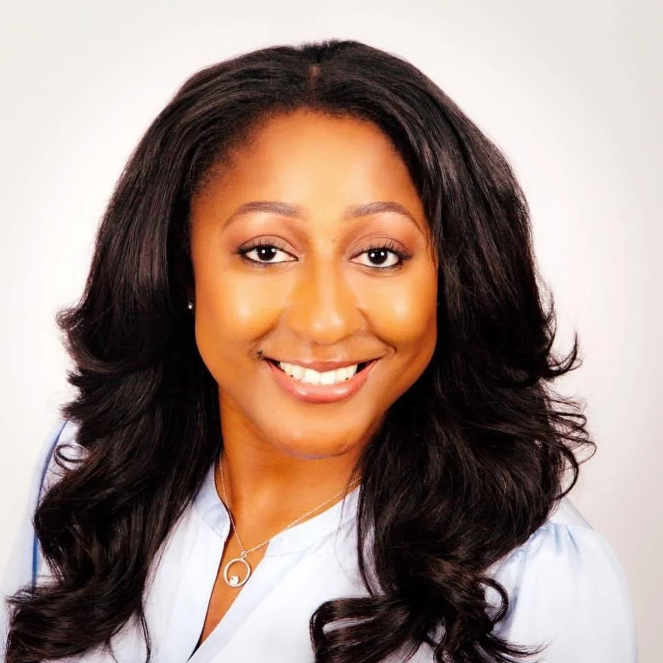 A woman with long, wavy black hair smiling, wearing a light-colored blouse and a necklace, against a plain background.