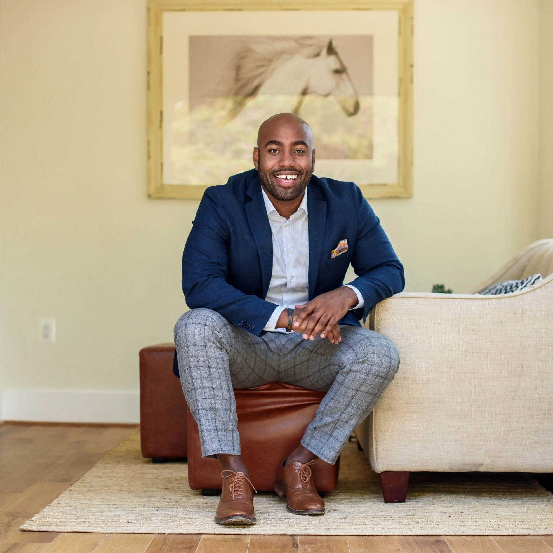 Man in a blue blazer and checked pants sitting on a brown ottoman in a living room with a framed picture of a white horse in the background.