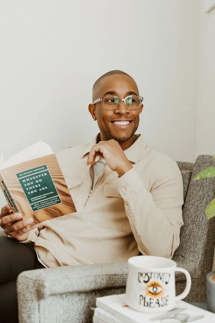 A smiling man with glasses sitting on a couch, holding a book. There is a white coffee mug with the words "Positivity Please" on it, placed on a stack of books in front of him.
