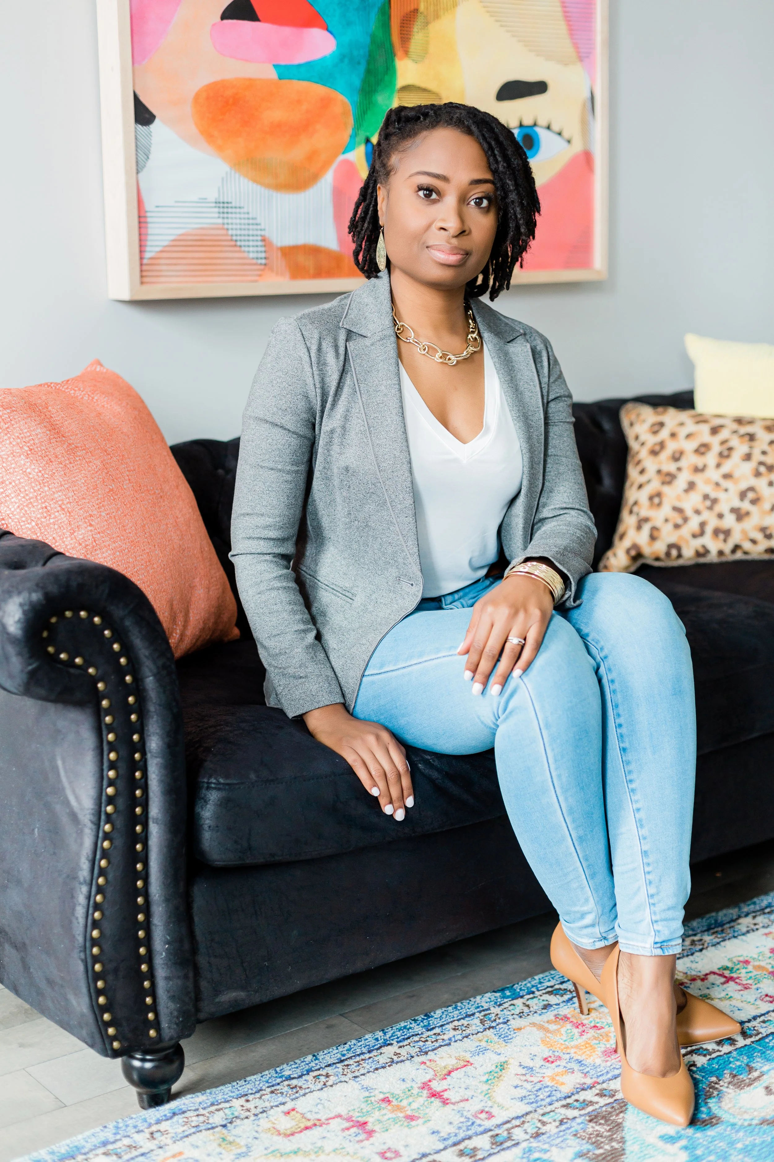 A woman with dark curly hair styled in dreadlocks, wearing a gray blazer, white top, light blue jeans, and tan high heels, sitting on a black velvet couch with colorful throw pillows and a framed abstract art piece in the background.