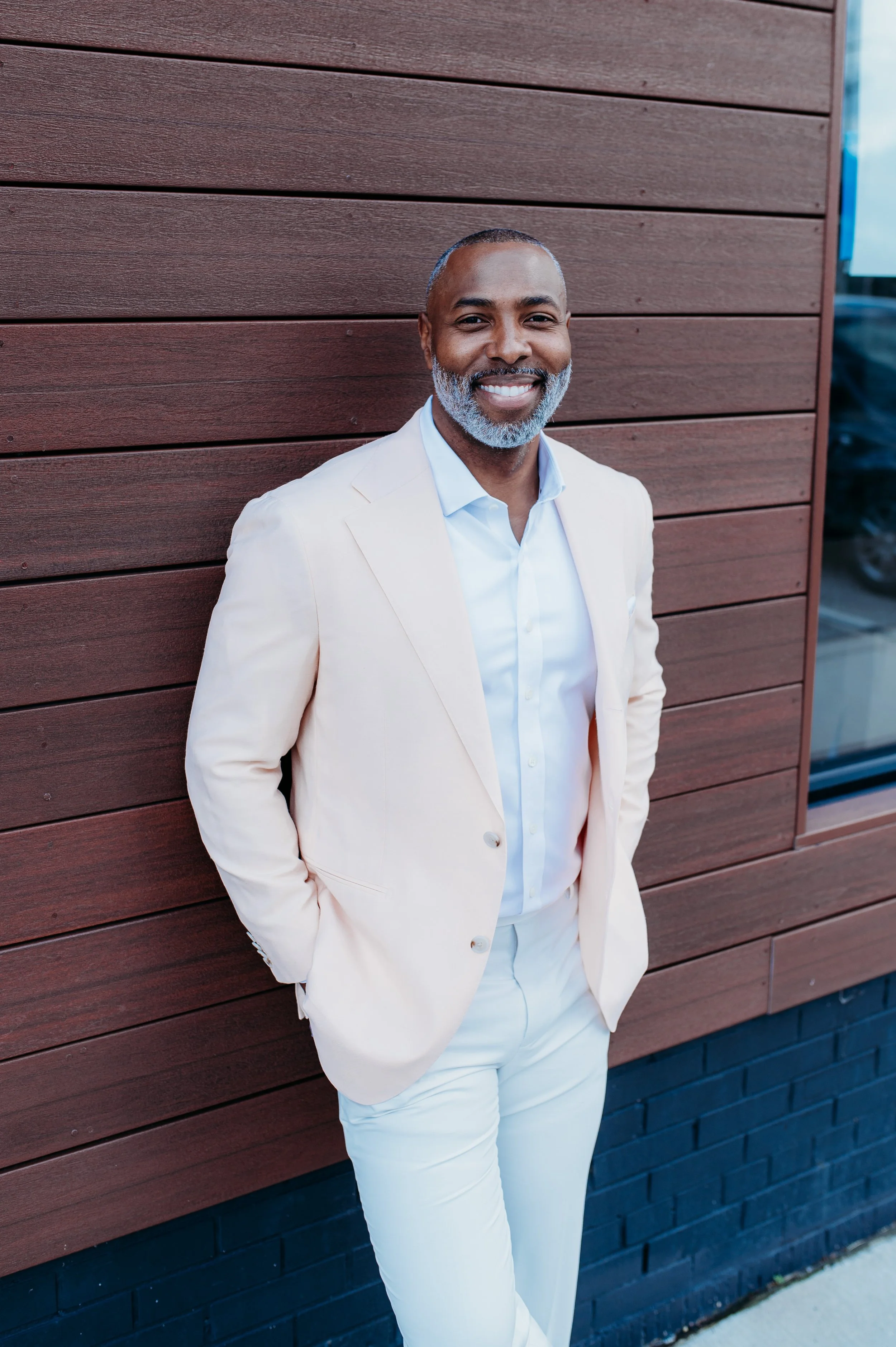 A smiling middle-aged man with grey beard, dressed in a white suit and shirt, leaning against a wooden wall outdoors.