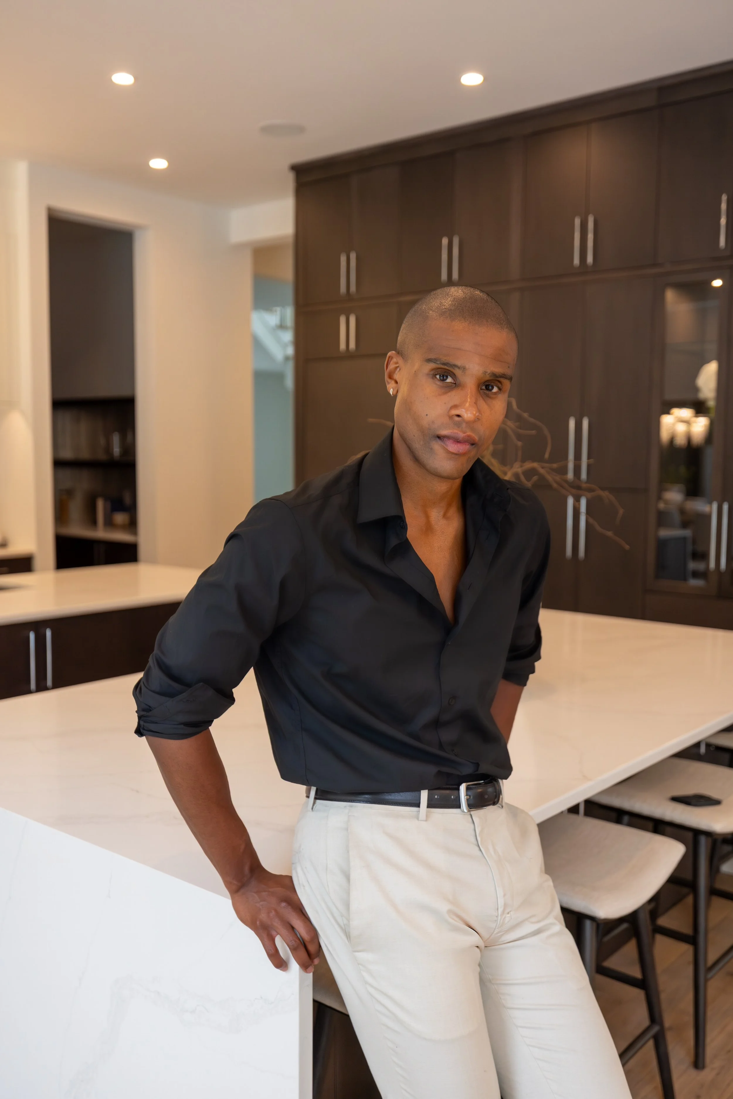 A man in a black shirt and beige pants leaning against a white kitchen island in a modern kitchen.