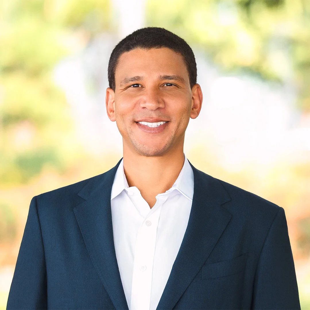 A smiling man in a suit and white shirt outdoors with blurred green leaves in the background.