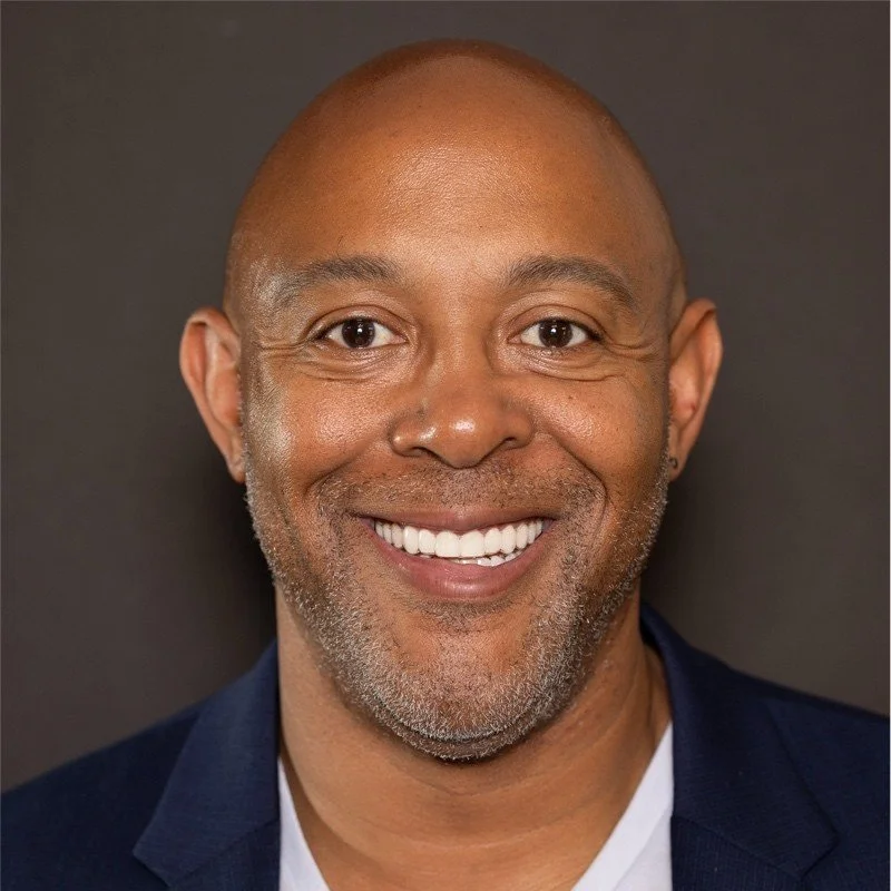 Close-up portrait of smiling bald man with a beard, wearing a dark blazer and white shirt, against a dark background.