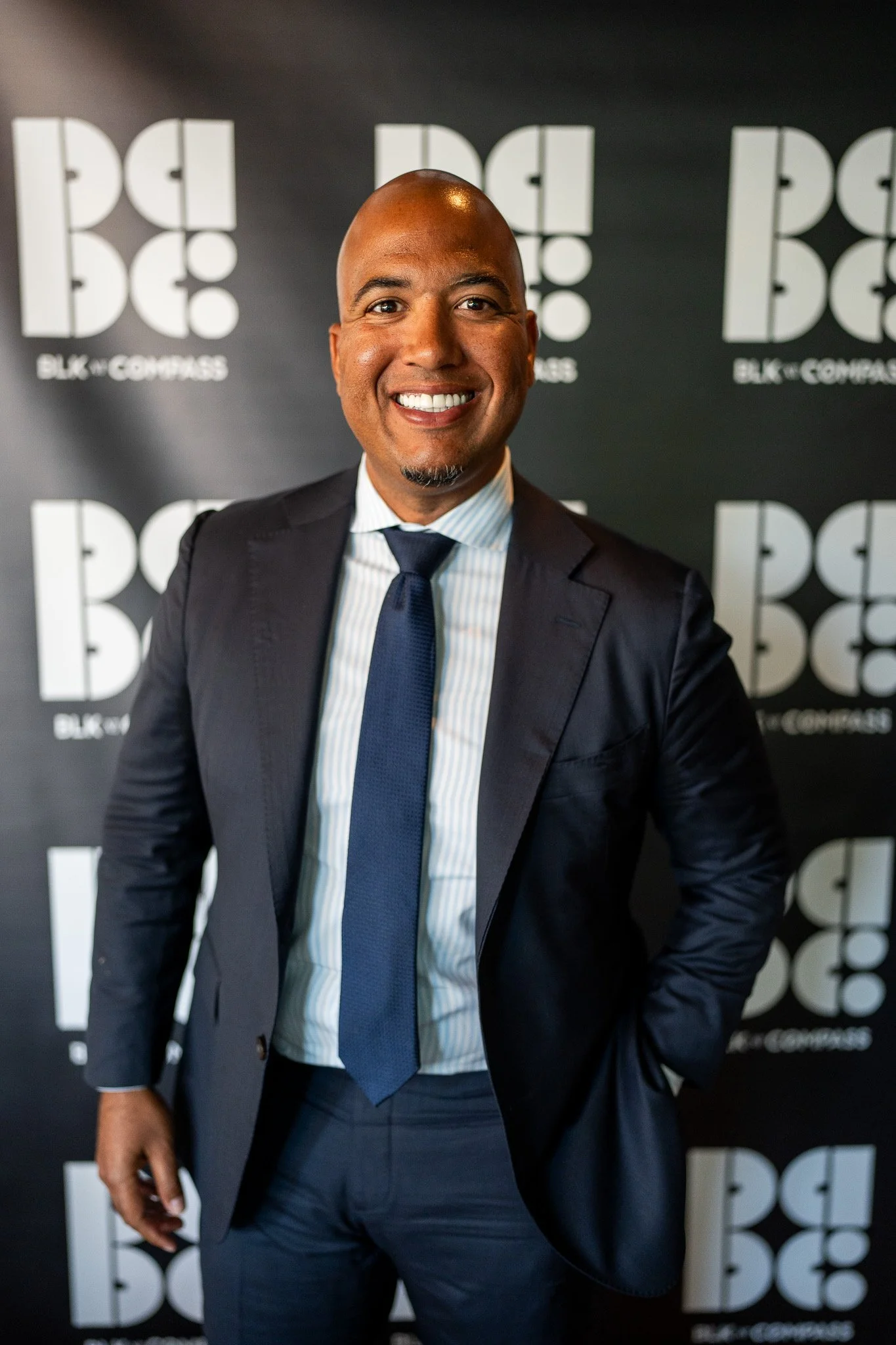 A smiling man in a dark suit, white dress shirt, and blue tie standing in front of a black backdrop with white logos that read 'BLK + CODE'.