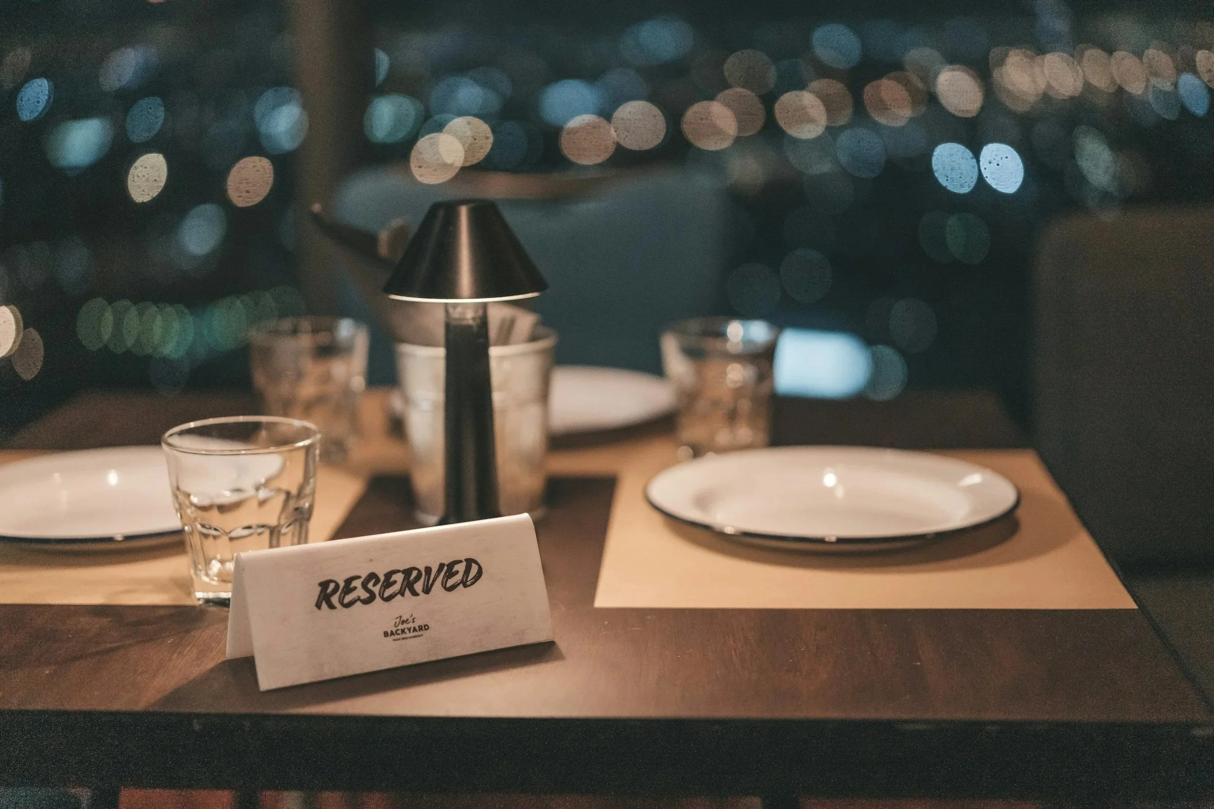A reserved table at a restaurant with empty plates, glasses, a small black lamp, and a 'Reserved' sign, with a cityscape view of blurred city lights in the background.
