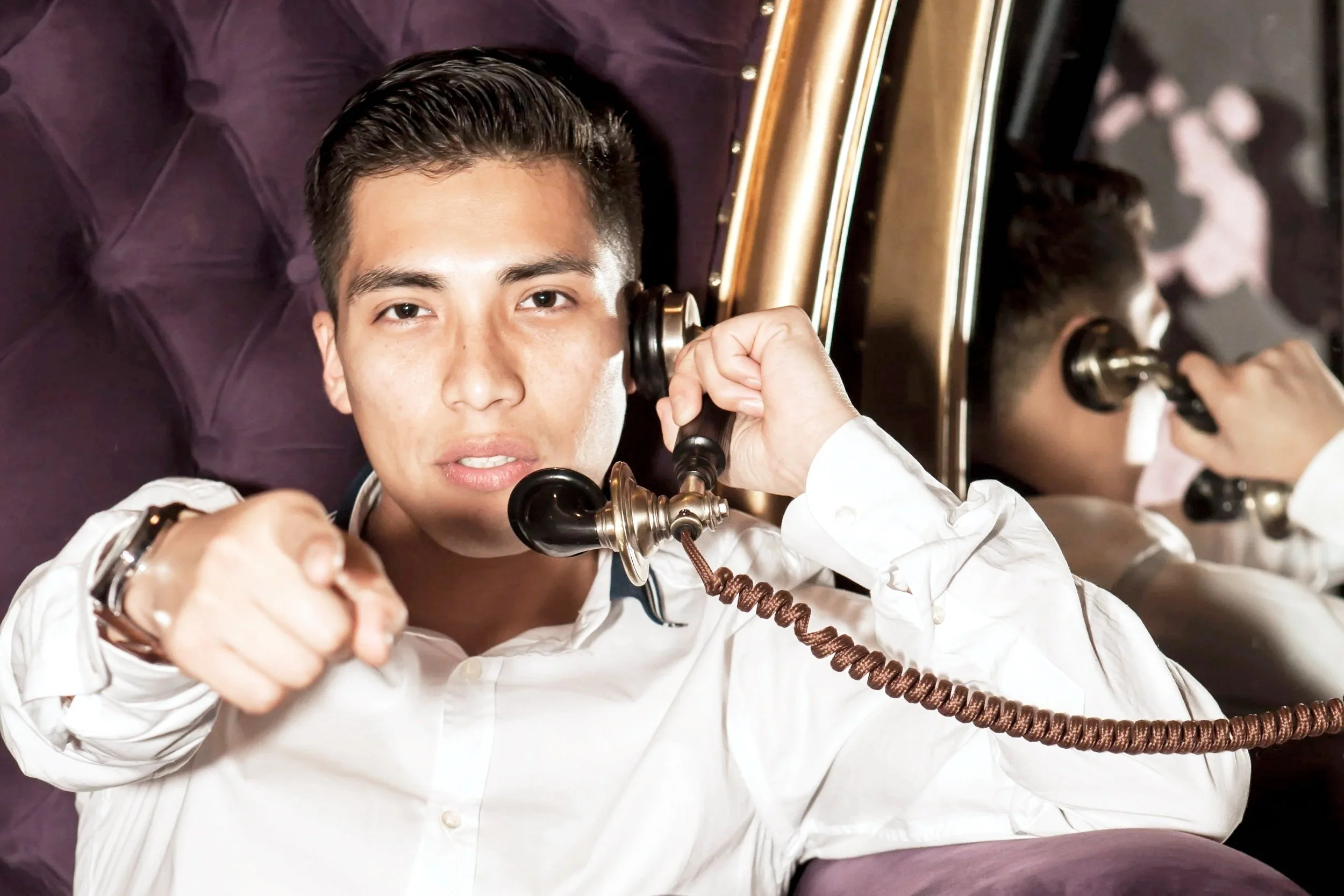 Young man with dark hair and light skin holding a vintage telephone receiver to his ear, sitting on a purple velvet headboard with mirrored accents, looking at the camera.