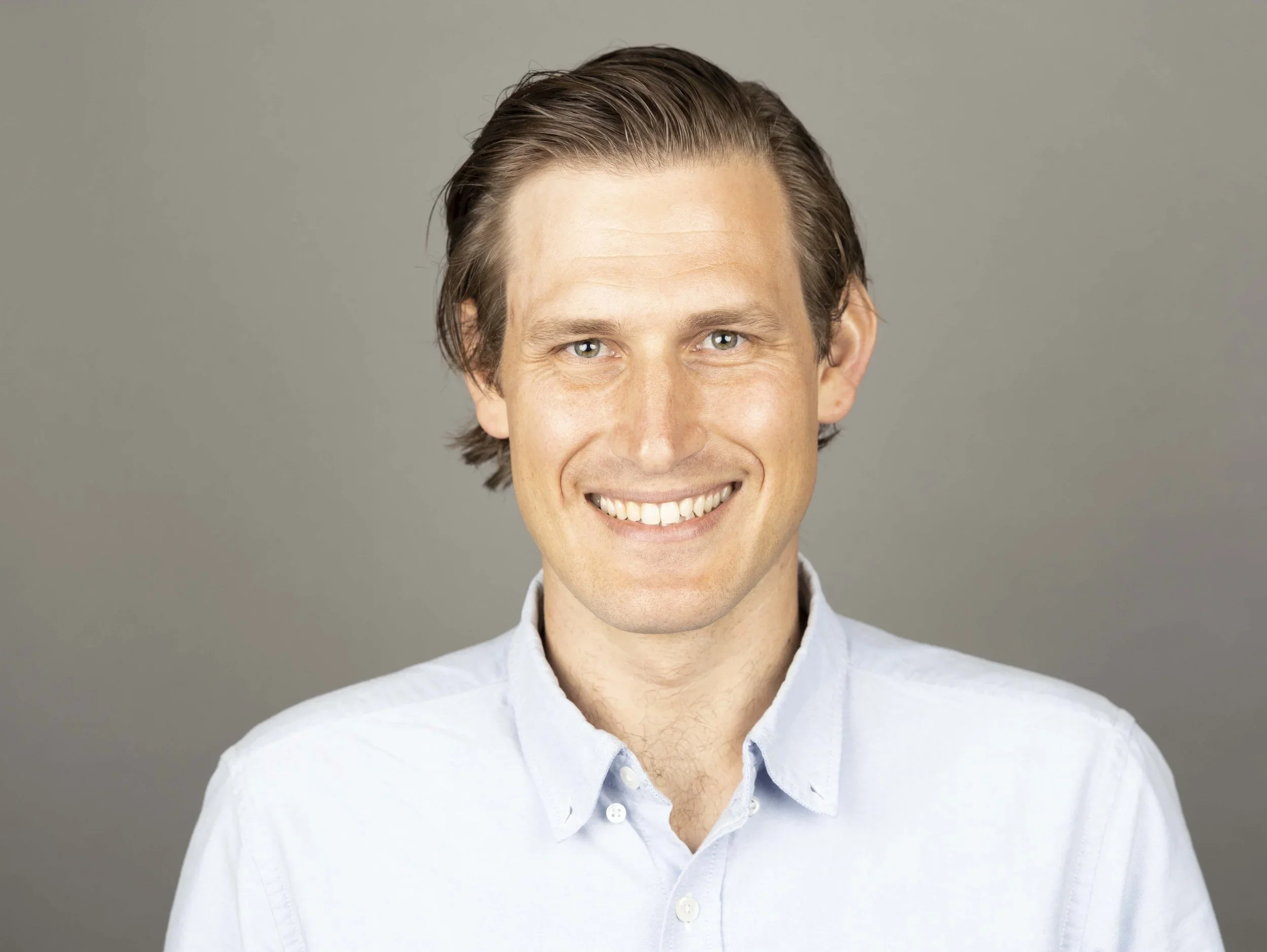 Portrait of a smiling man with brown hair and blue eyes, wearing a light blue button-up shirt, against a plain gray background.