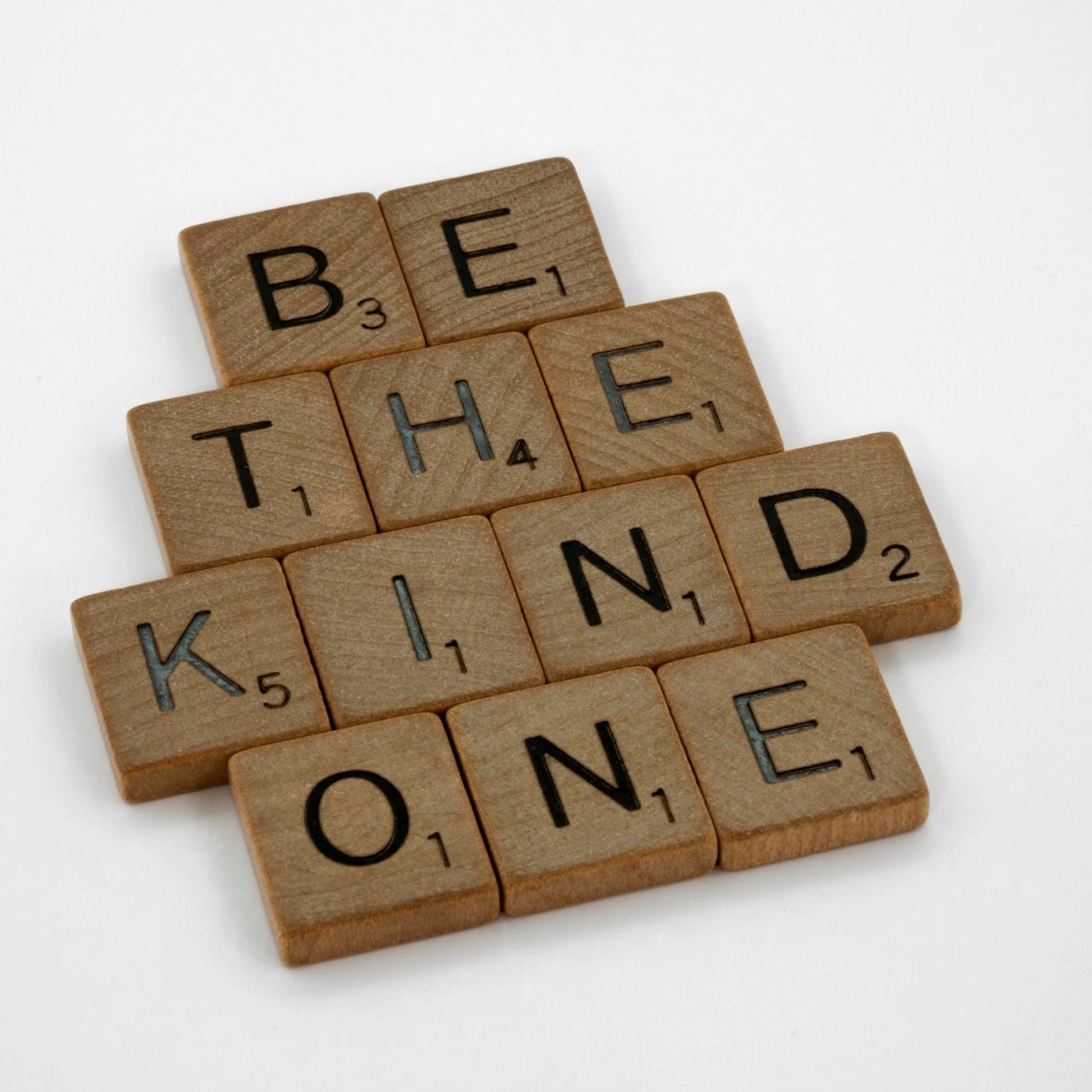 Wooden Scrabble tiles spell out 'BE THE KIND' on a white background.