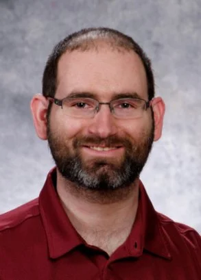 Headshot of a man with glasses, a beard, and short hair, wearing a maroon collared shirt, smiling against a gray background.
