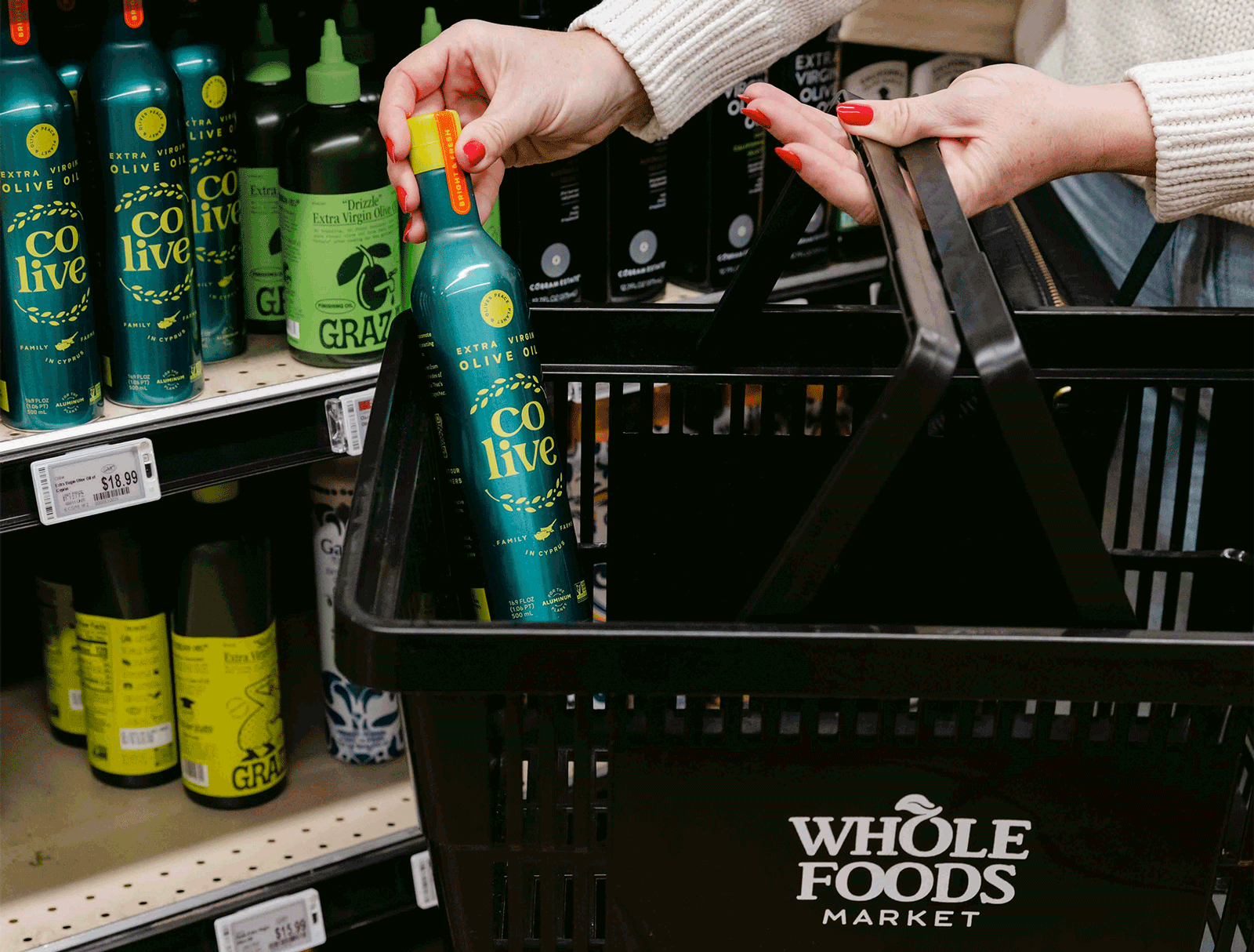 Person placing a bottle of Co Live Extra Virgin Olive Oil into a Whole Foods Market shopping basket, with more bottles displayed on a store shelf.