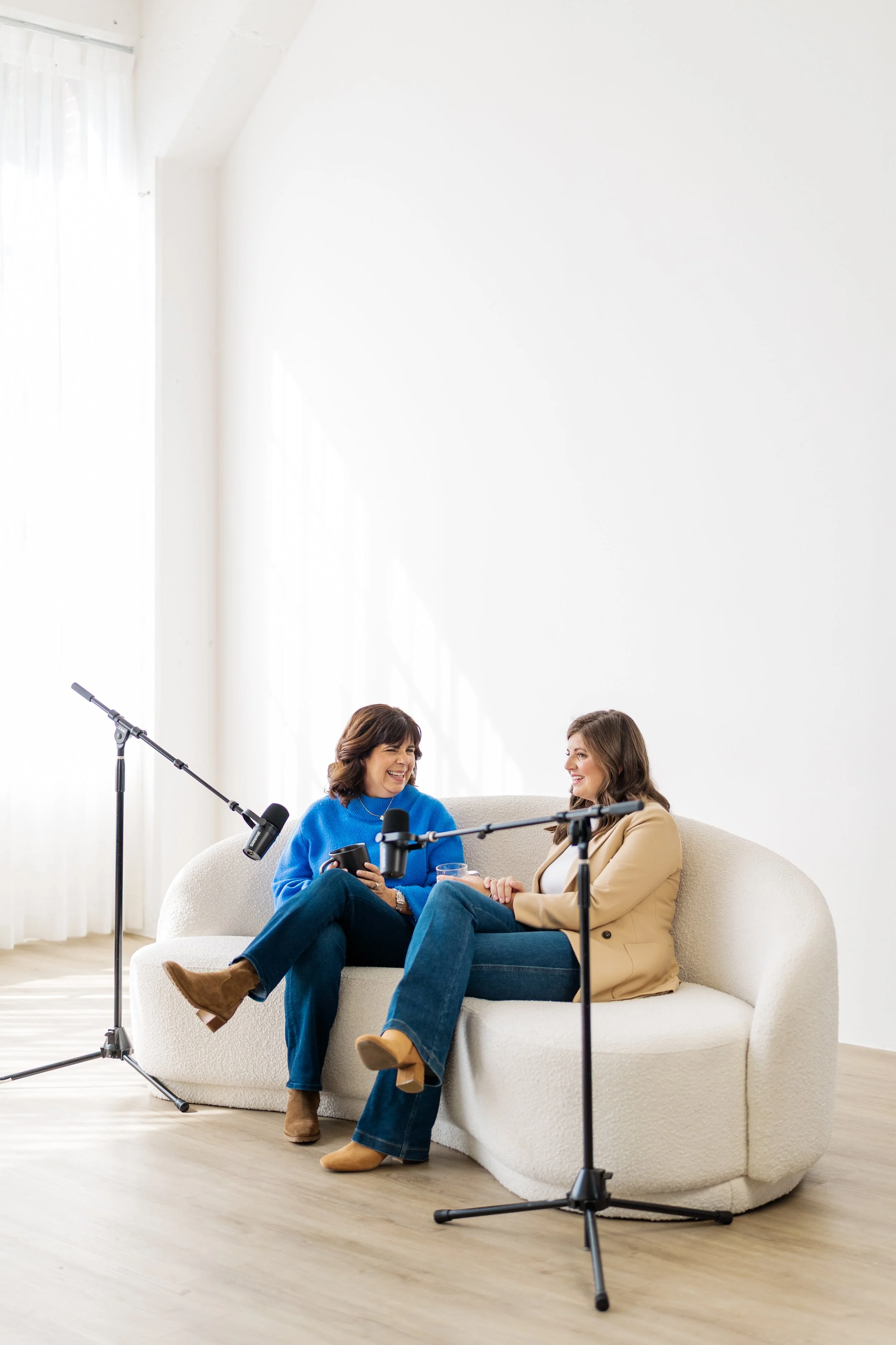 Two women are seated on a white sofa engaged in conversation, in front of microphones.
