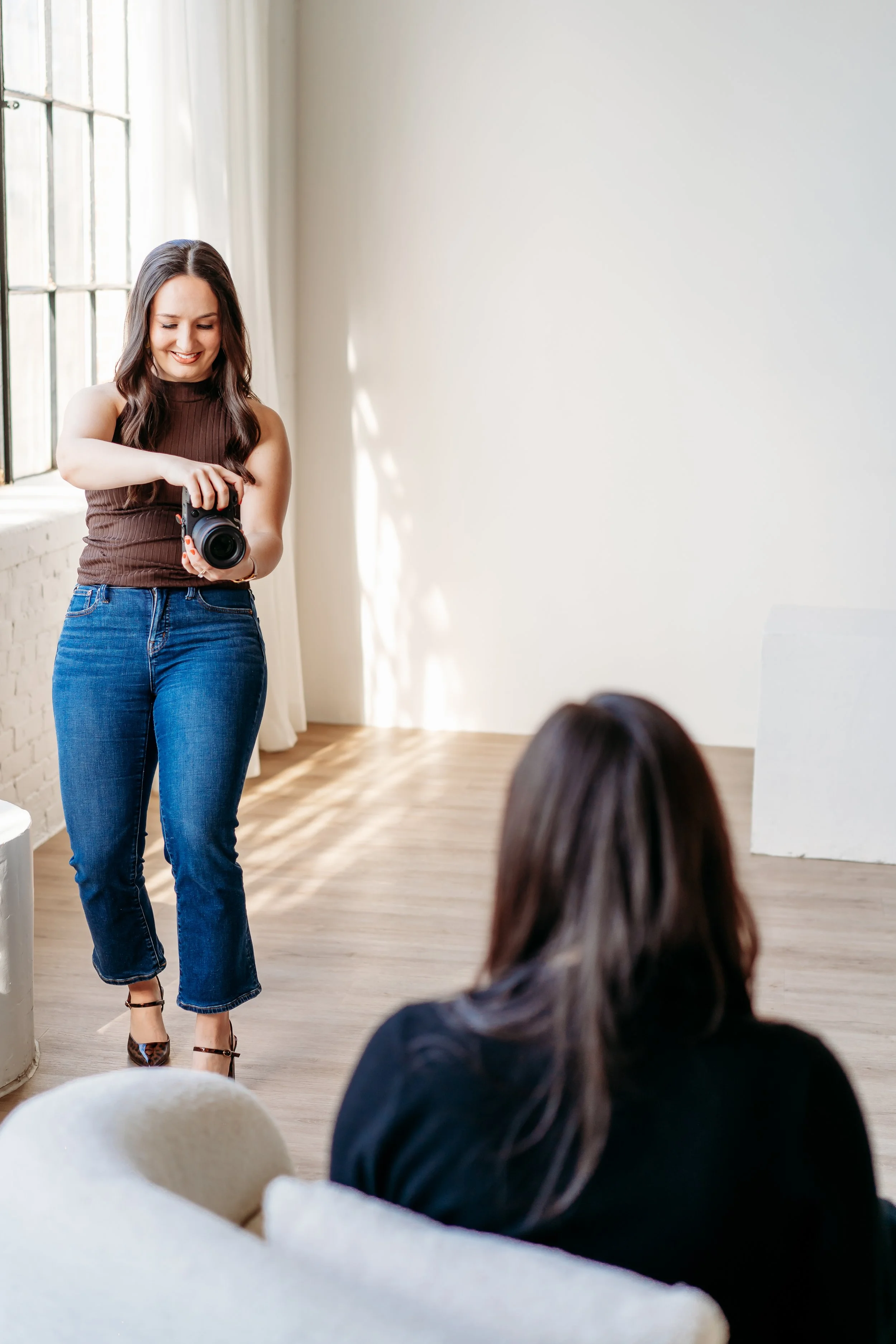 A woman stands in front of a bright window smiling as she takes a photo of another woman that is sitting in the foreground.