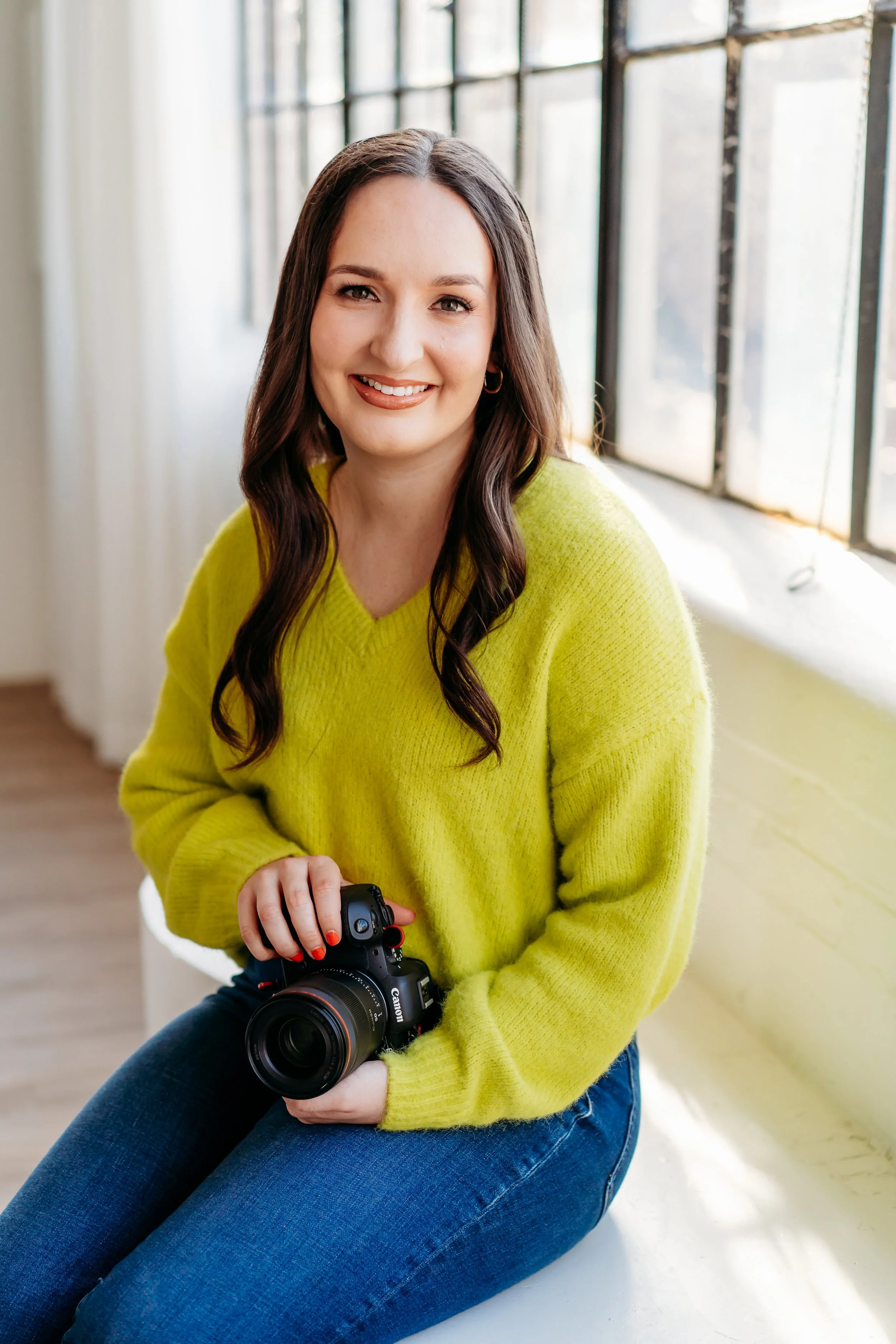 A woman with long brown hair in a bright sweater holds a camera, while posing for a picture.