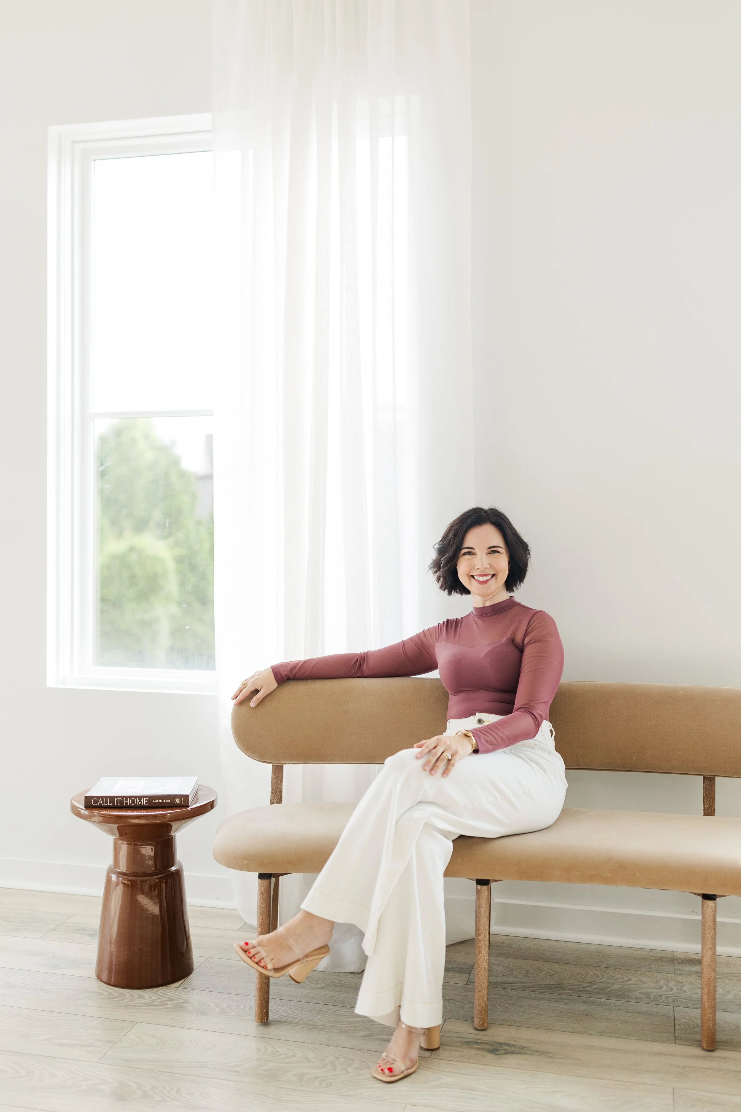 A woman in white pants and a purple top sitting on a tan bench by a window.