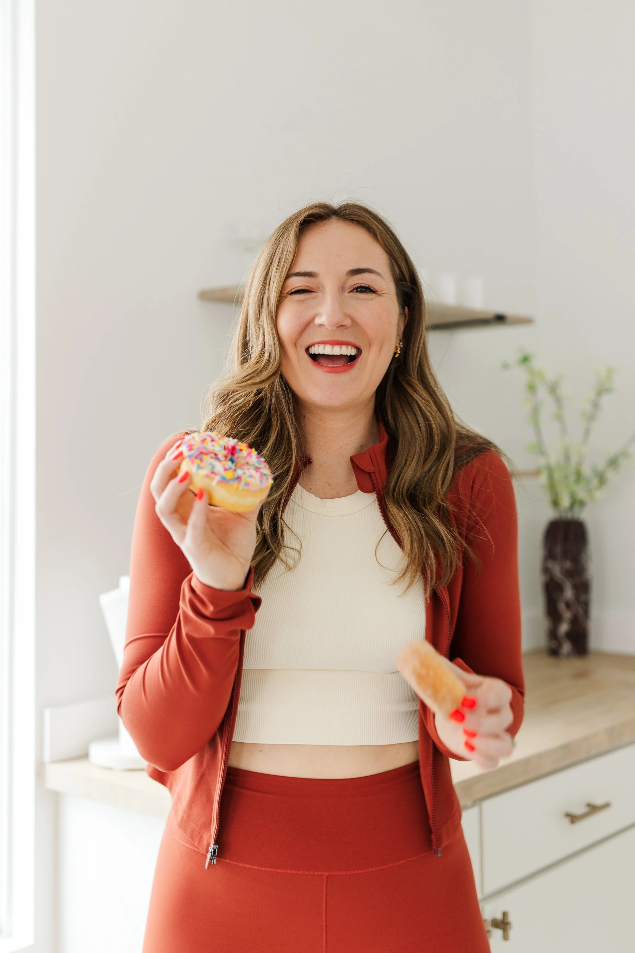 A woman joyfully holds a sprinkle-covered donut, wearing a red jacket and white top.