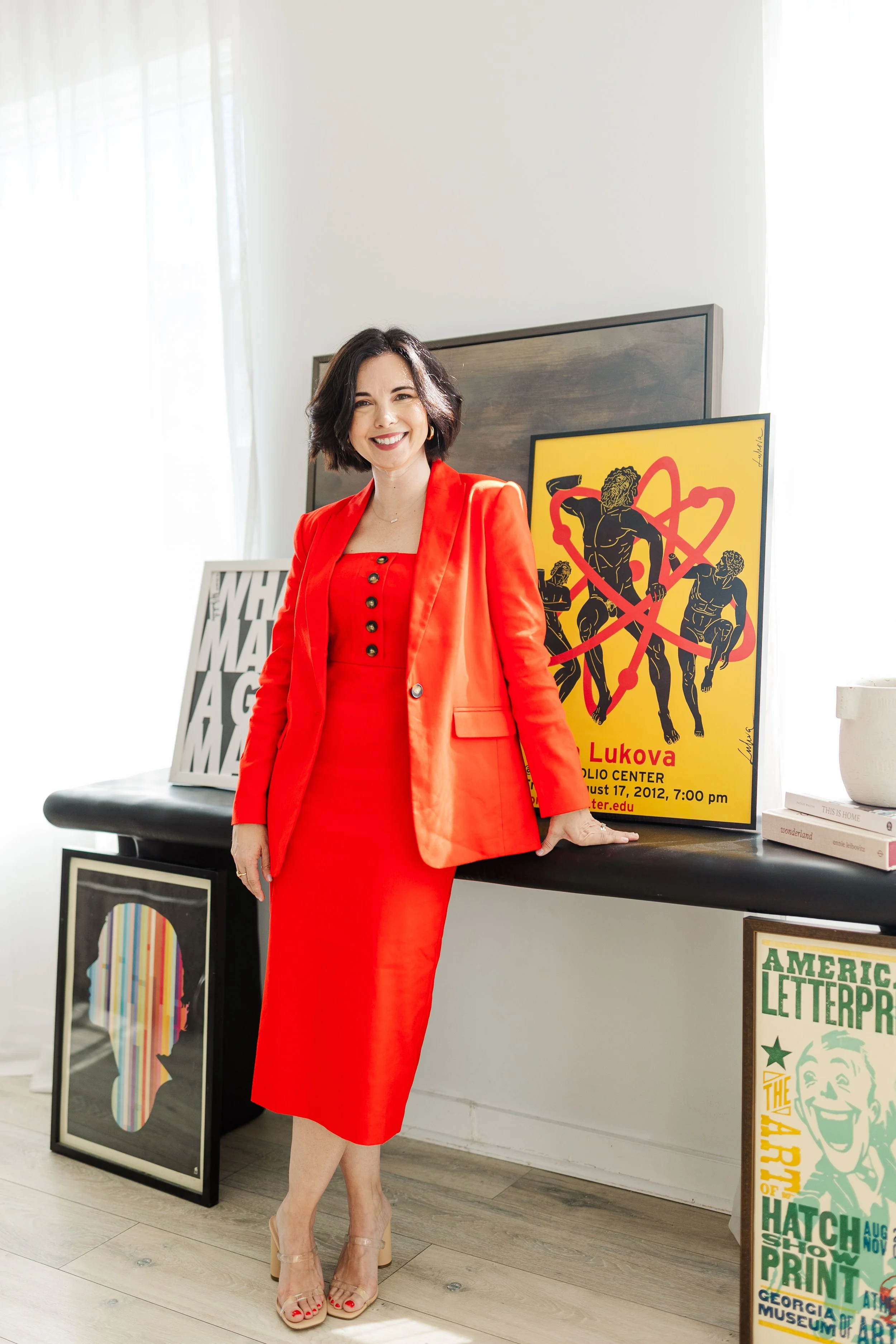 A woman in a bright red outfit smiles, standing beside an art table.
