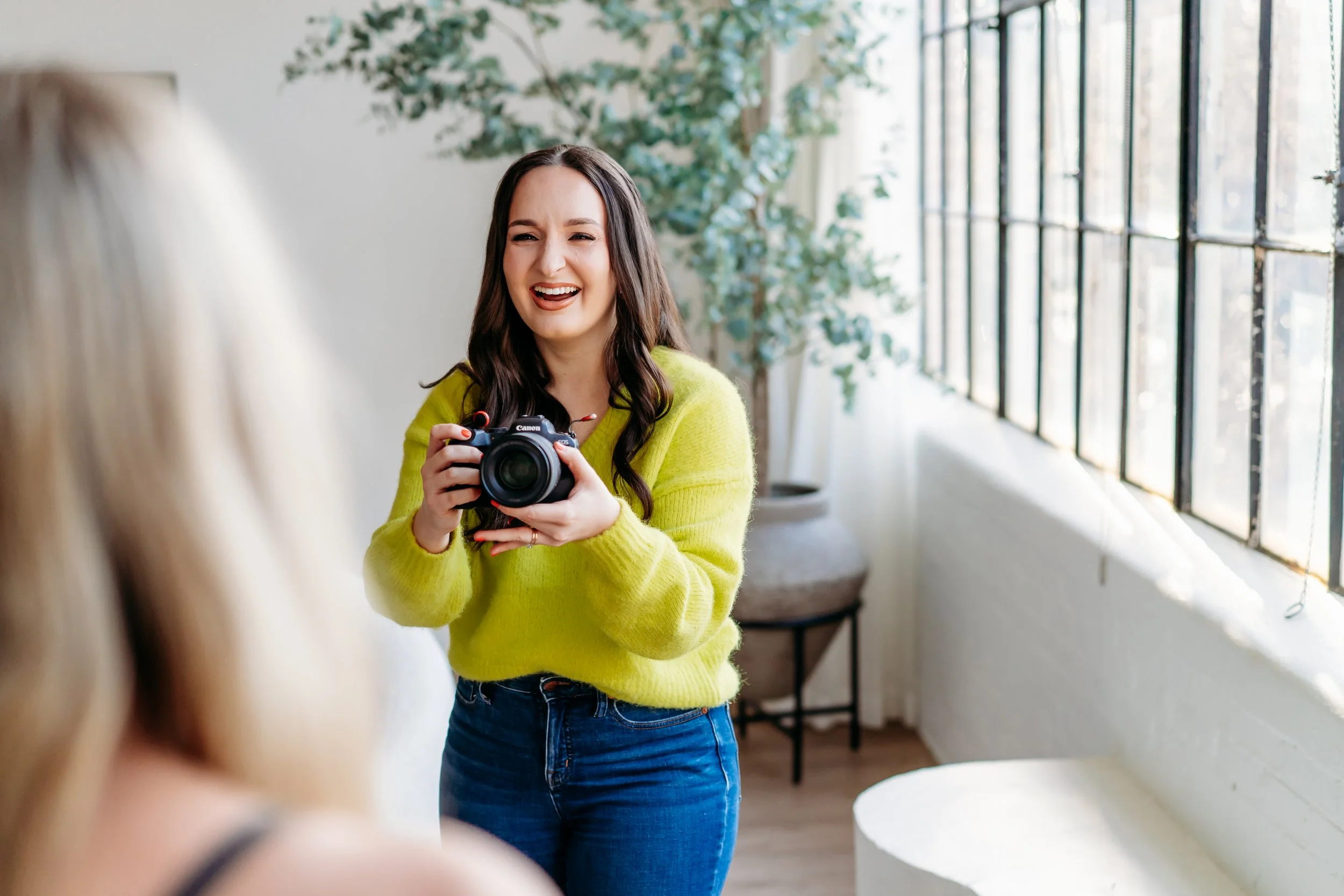 A woman in a bright sweater smiles joyfully while holding a camera.
