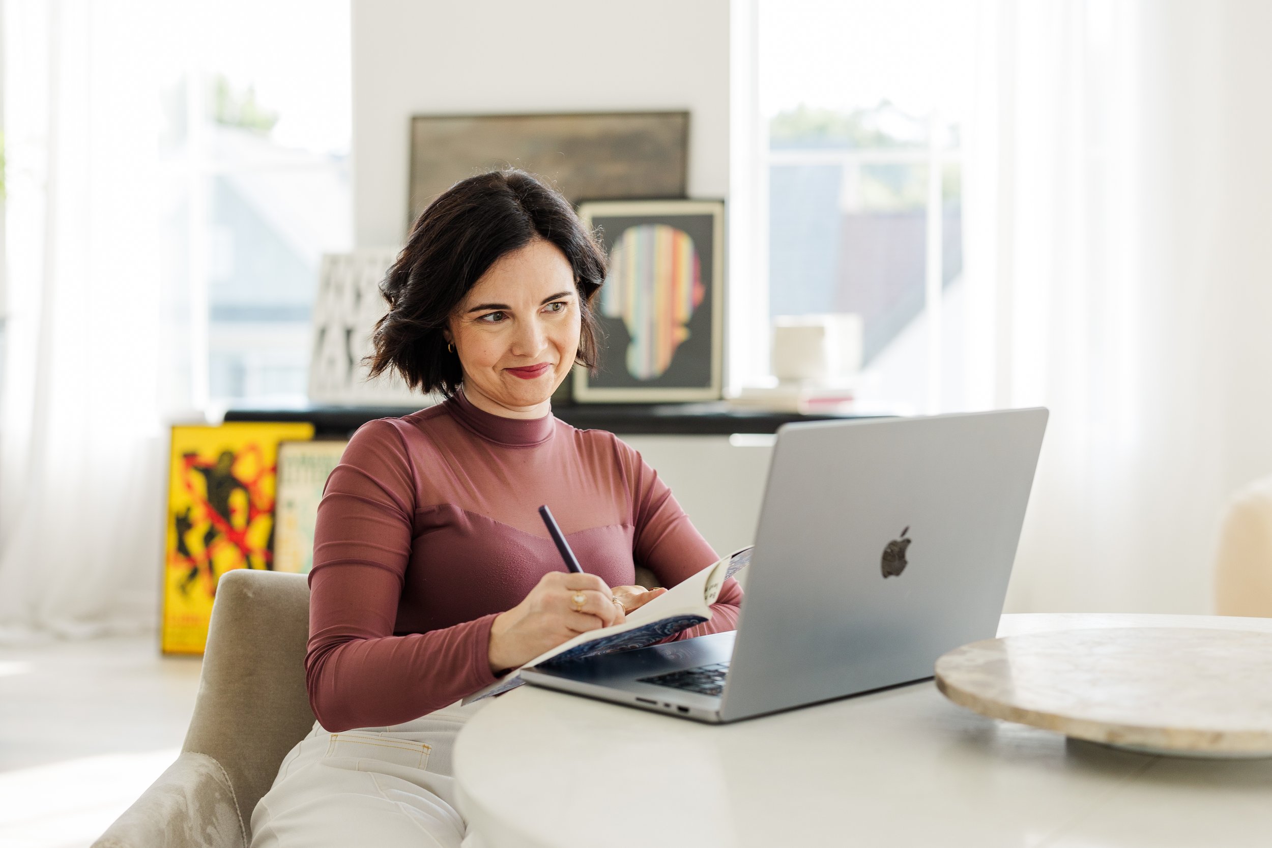 A woman in a maroon turtleneck sits at a table, writing in a notebook beside a laptop.