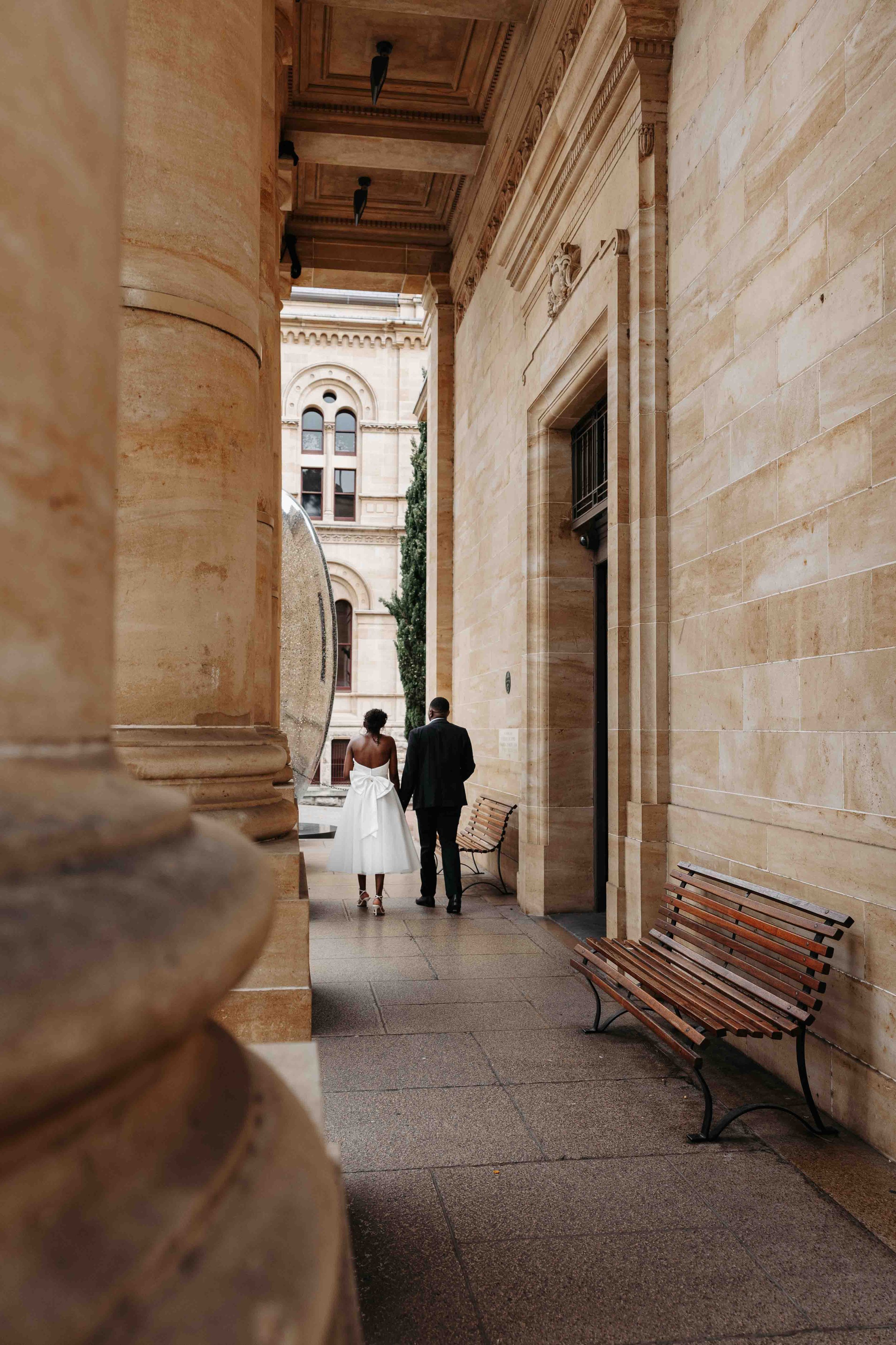 Newly weds walk hand in hand under the porch of the Adelaide Art Gallery. The image has natural colours. Captured by Adelaide Wedding Photographer, Woodley Photography.