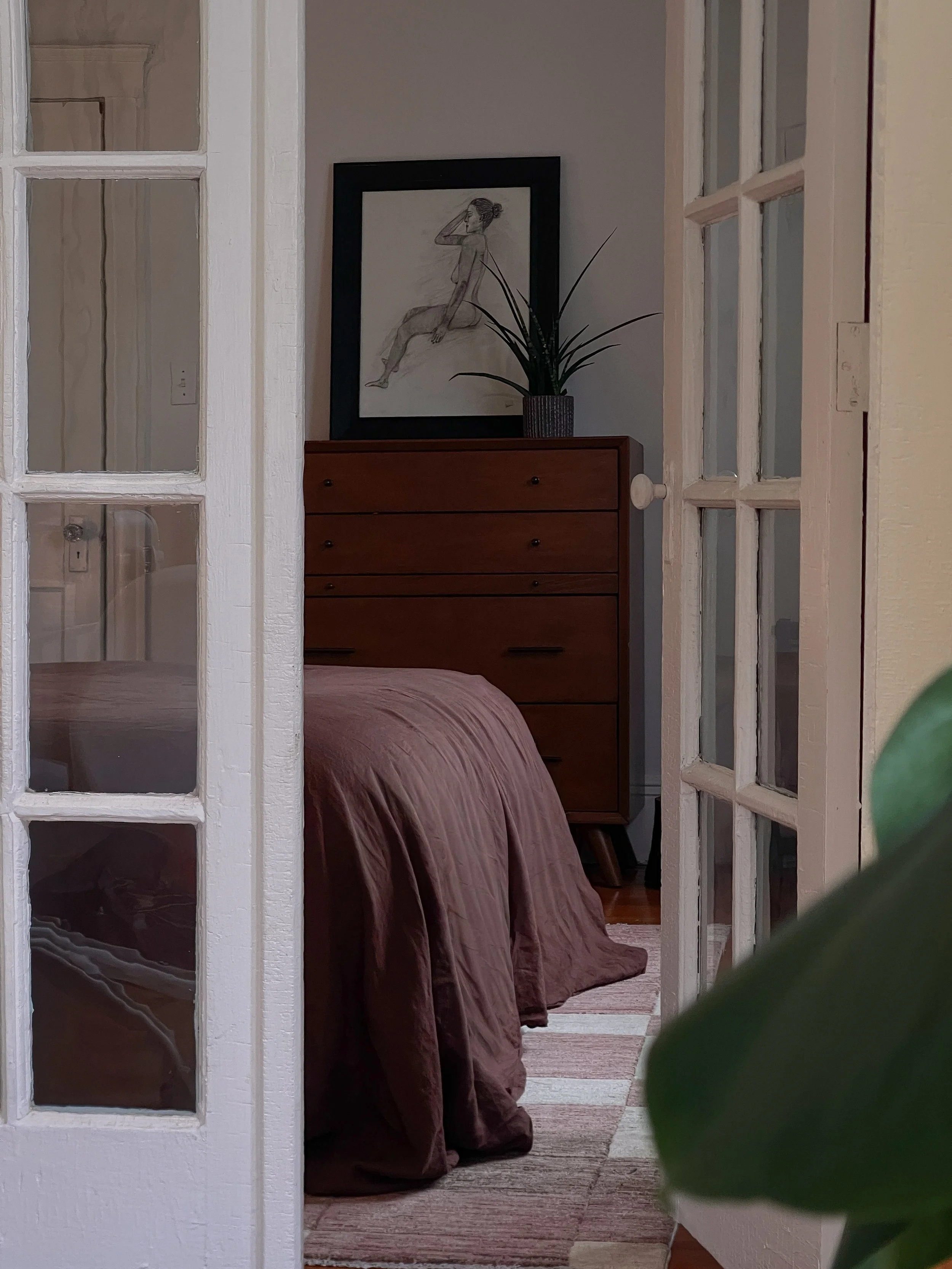 Photograph of a bedroom viewed through French doors, featuring a mahogany dresser, linen bedding in dusty rose, and a figure drawing in a black frame.
