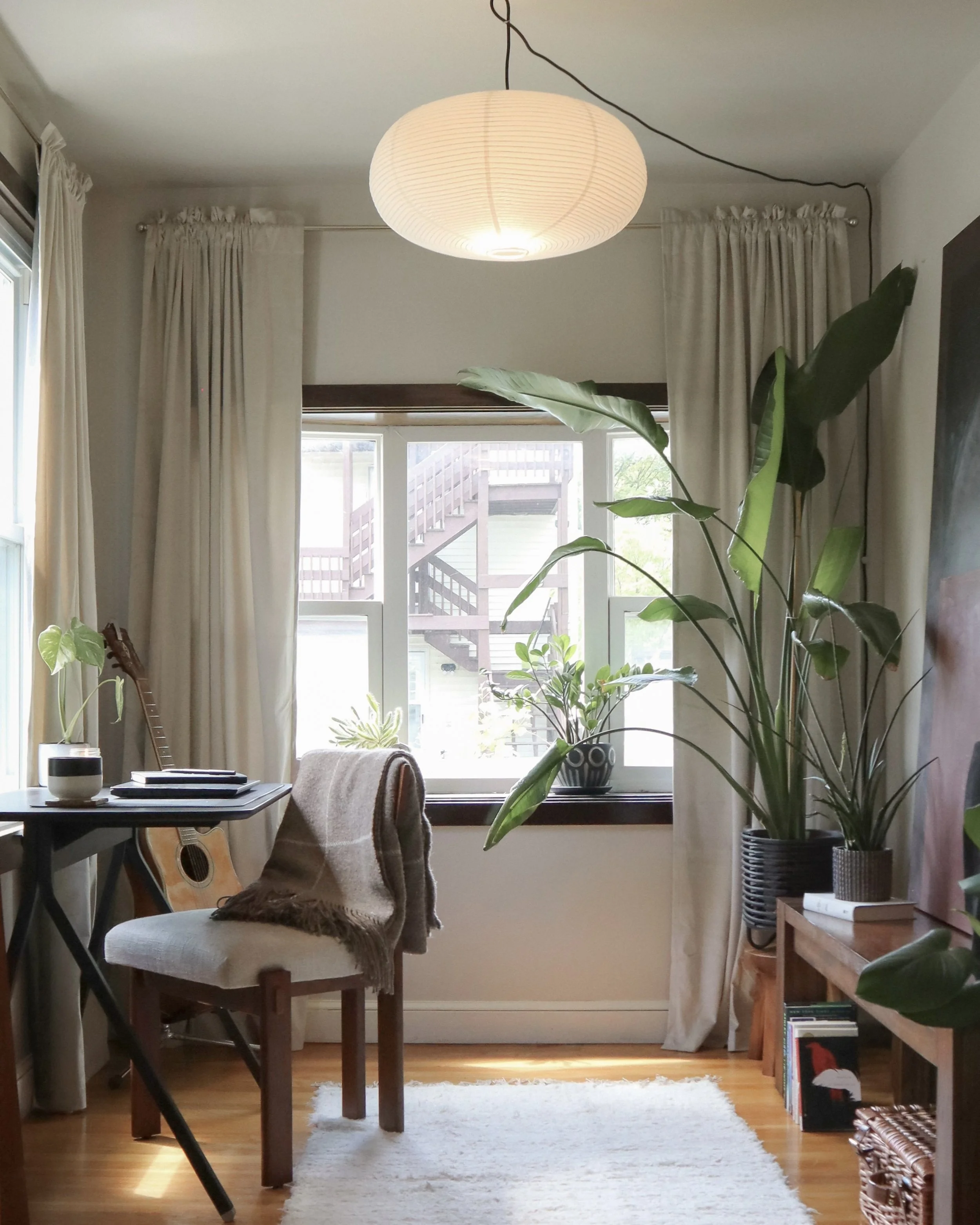 Photograph of a light-filled home office with linen curtains, a Noguchi-style pendant light, a guitar leaning against a desk, and a large bird of paradise plant.