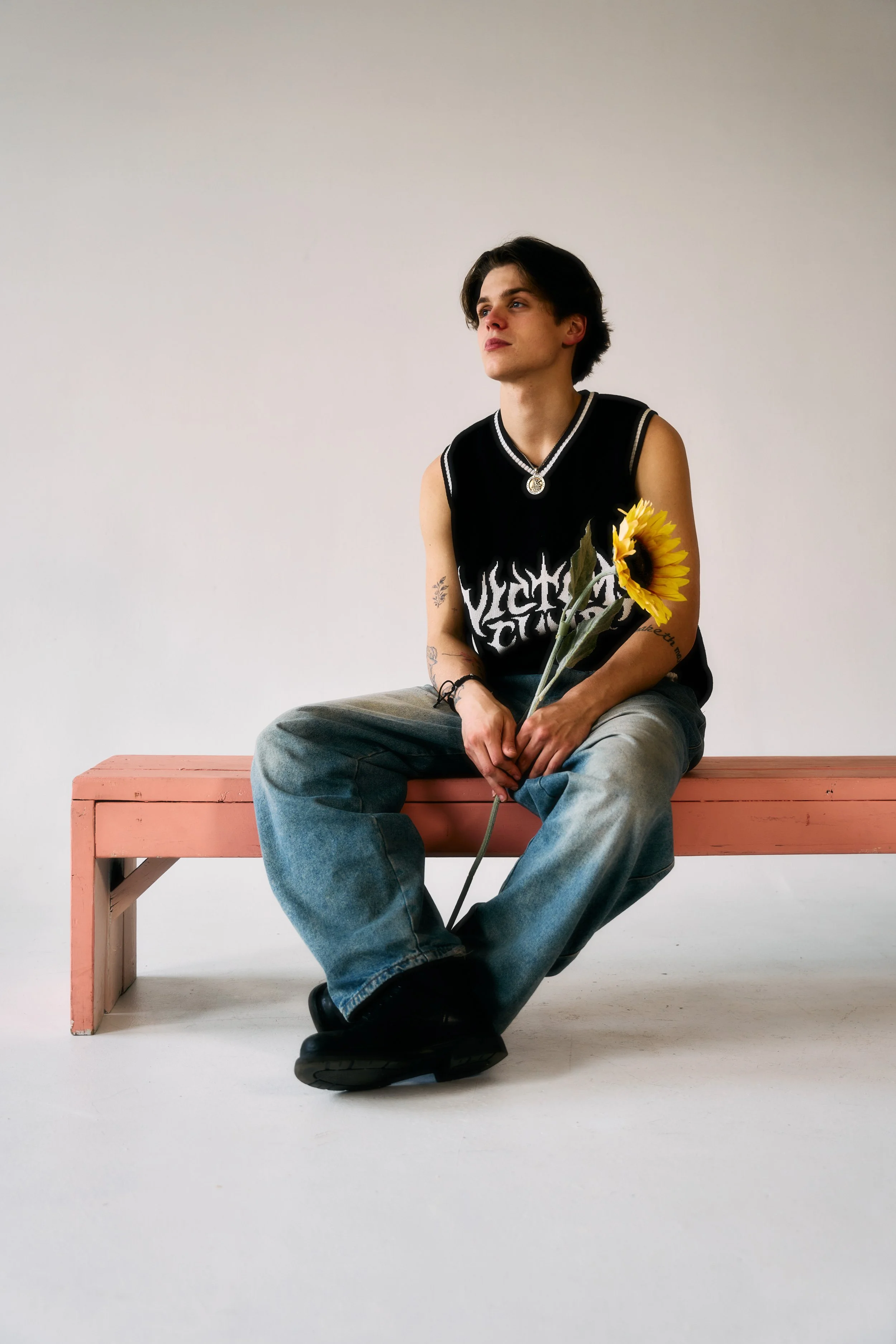 A young man with dark, wavy hair sitting on a wooden chair against a plain, light gray background. He is dressed in a dark sweatshirt with a graphic design, oversized dark green cargo pants, and sneakers. His posture is relaxed, with one leg crossed over the other, and he appears to be gazing forward.